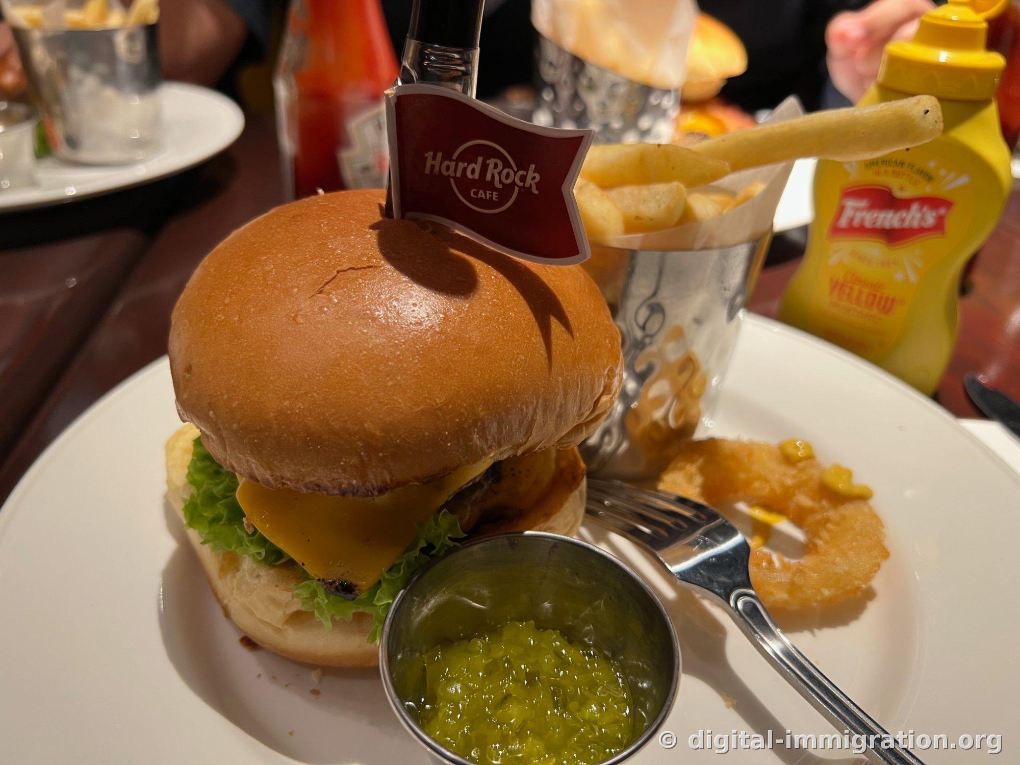 Hard Rock Cafe 로고 깃발이 꽂힌 나이프가 꽂힌 치즈버거와 금속 컵에 담긴 감자튀김, 초록색 피클 렐리시, 노란 프렌치스 머스터드 병