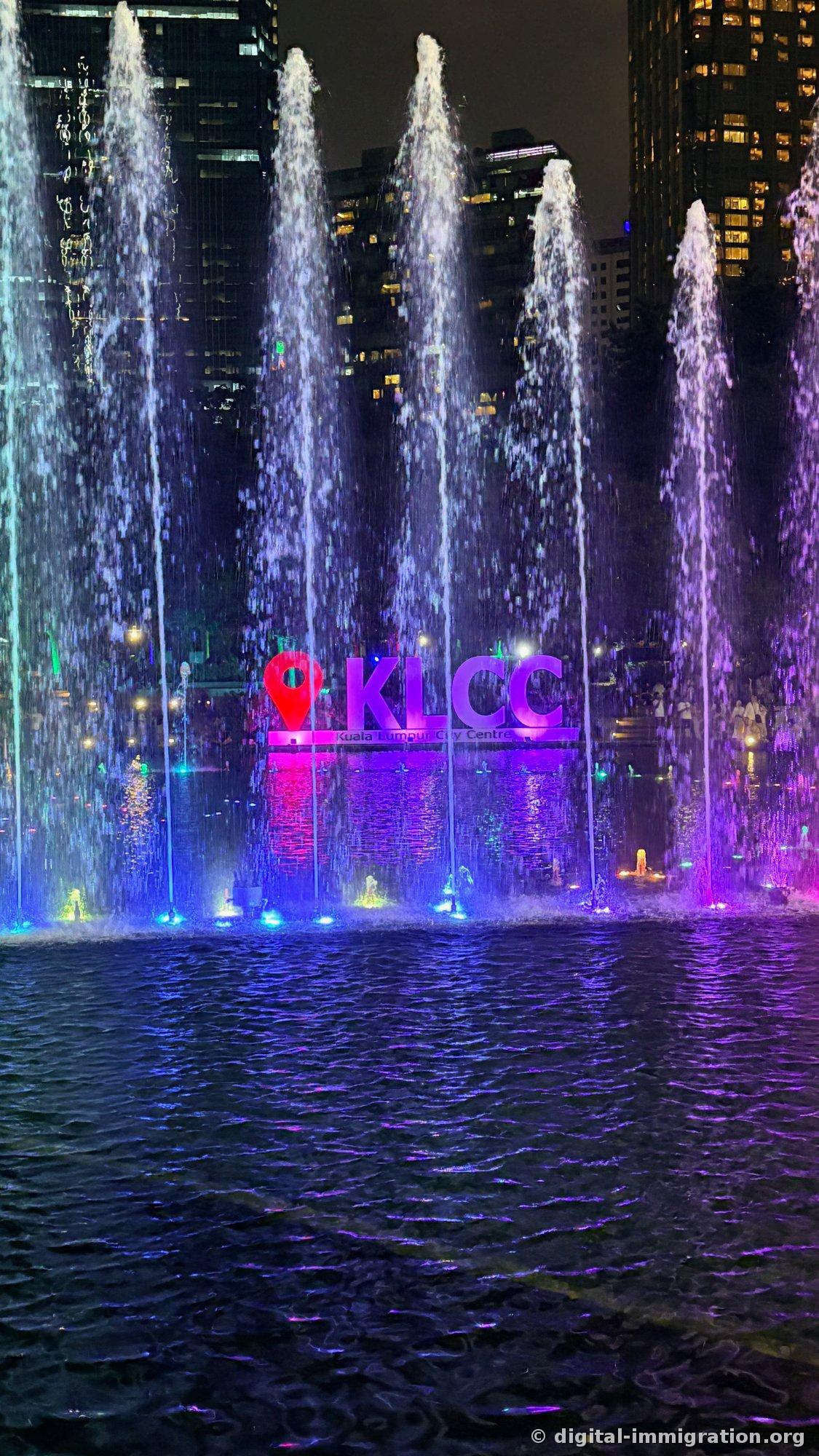 The Symphony Lake fountain show at KLCC at night. Water jets shot up high under purple and blue lighting, with the KLCC sign featuring a red pin icon visible between the jets and surrounding tall buildings lit in the background.
