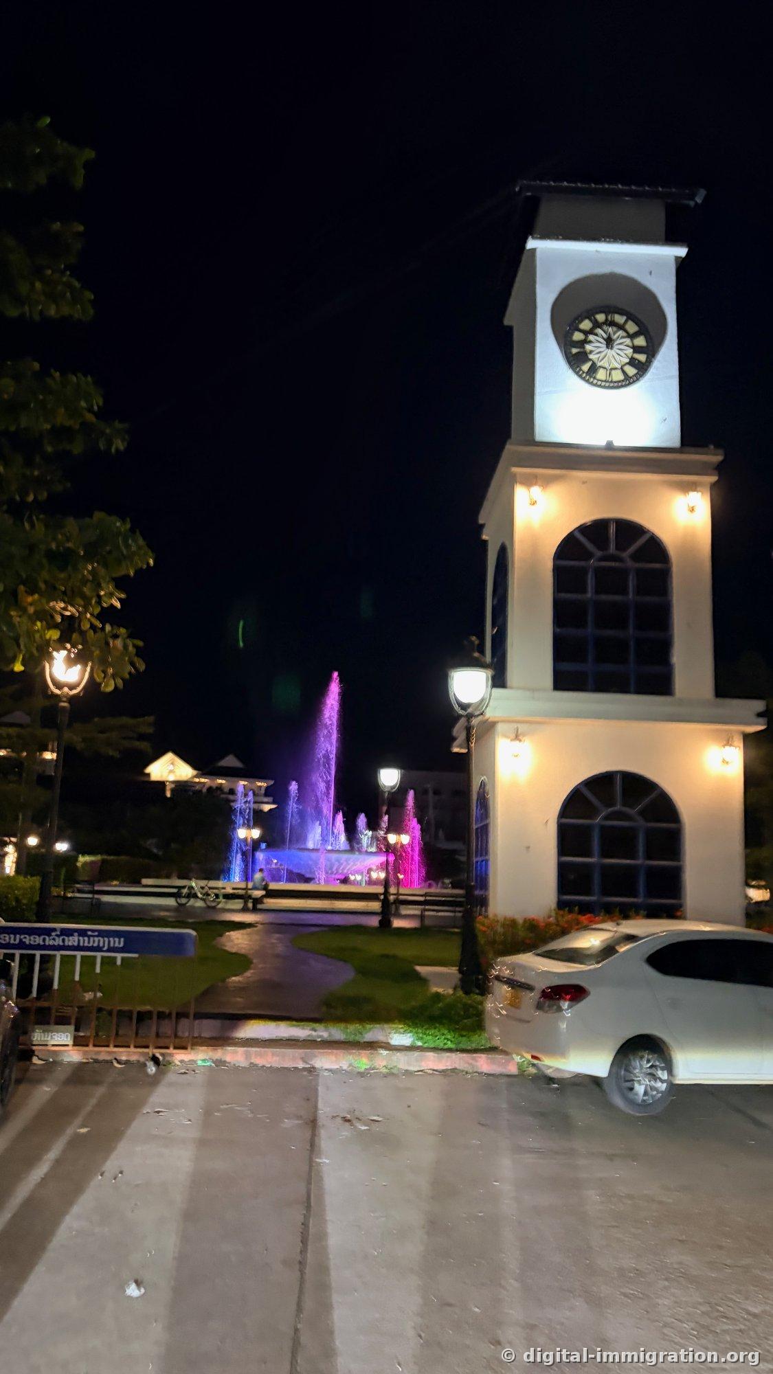 La tour horloge blanche illuminée et brillante la nuit, avec la fontaine Nam Phou teintée de rose et bleu au-delà, et une voiture blanche garée devant