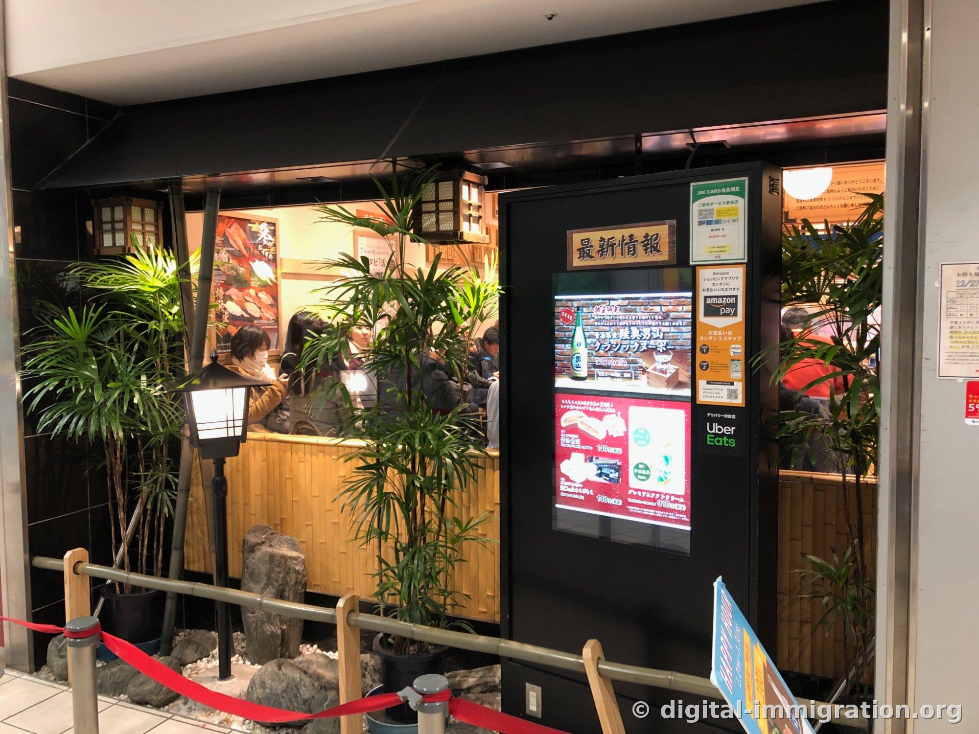 用竹子和景观石装饰的店前等候区，红色挡杆和「最新情報」数字标牌屏幕可见