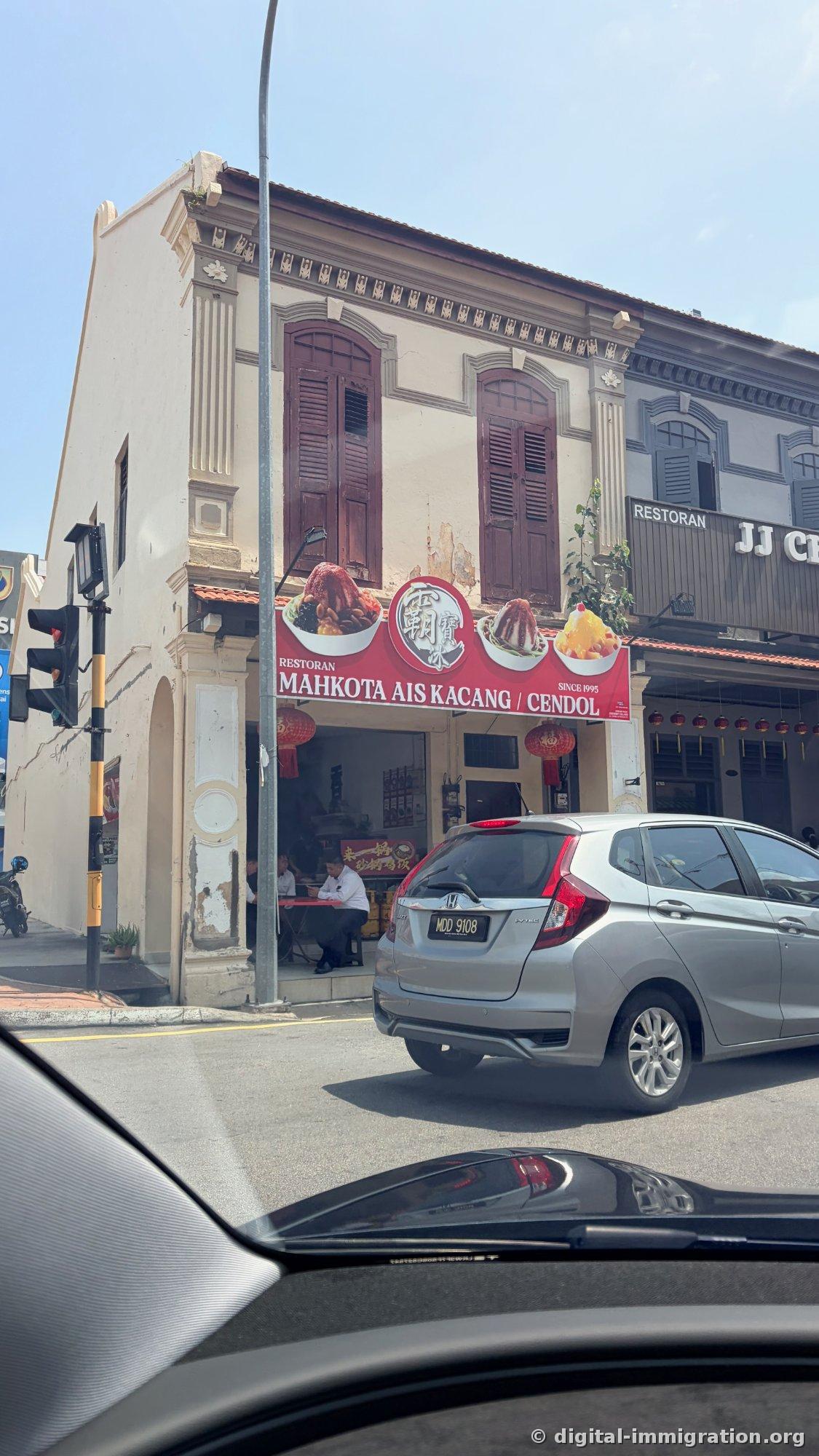 Storefront of a corner shophouse in Melaka shot from inside a car through the window. A red banner reading 'RESTORAN MAHKOTA AIS KACANG / CENDOL SINCE 1995' with three photos of shaved ice, brown wooden shutters, and red lanterns are visible, with a silver Honda car passing in front.