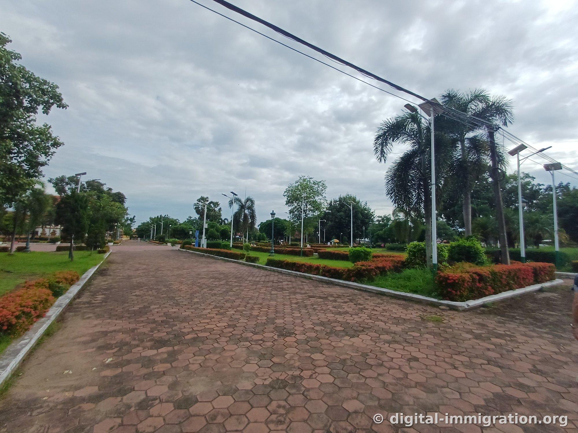 Red brick promenade lined with orange-hued shrub beds and palm-tree-shaped street lamps under a cloudy sky
