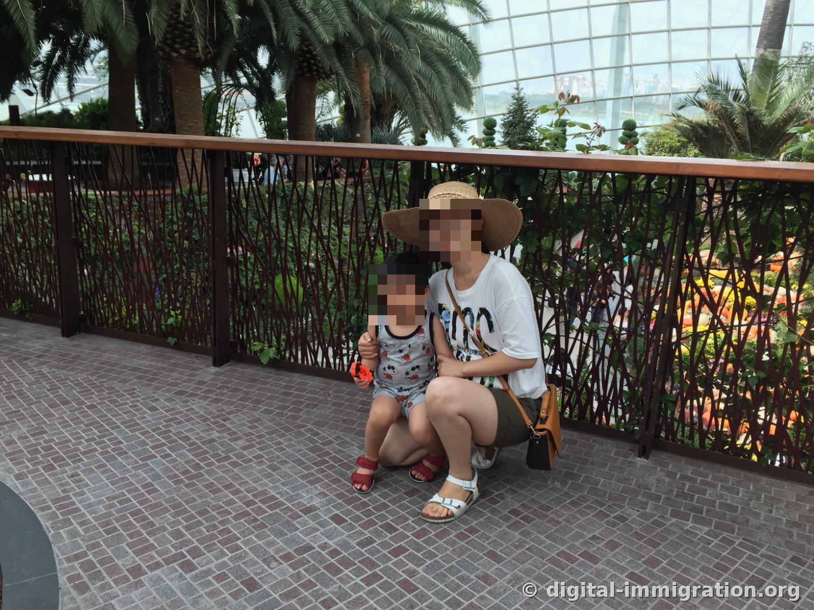 An adult and child sitting in front of the Flower Dome railing, with palm trees and the sea beyond the glass wall in the background
