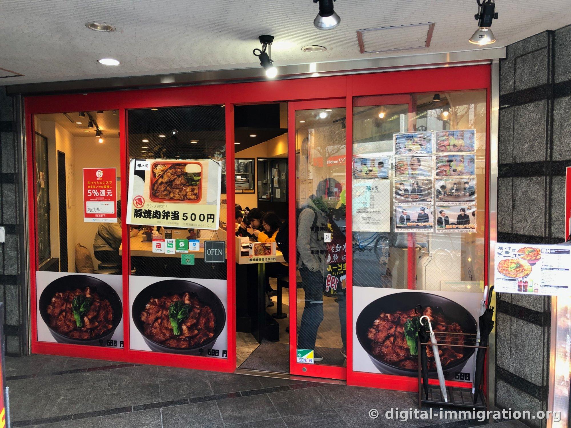 L'entrée de la succursale Shinjuku de Butadaigaku encadrée de rouge. La vitre affiche une photo de bol de porc et des panneaux indiquant « 中 600円 » et « 豚焼肉弁当 500円 », et des clients sont assis au comptoir à l'intérieur