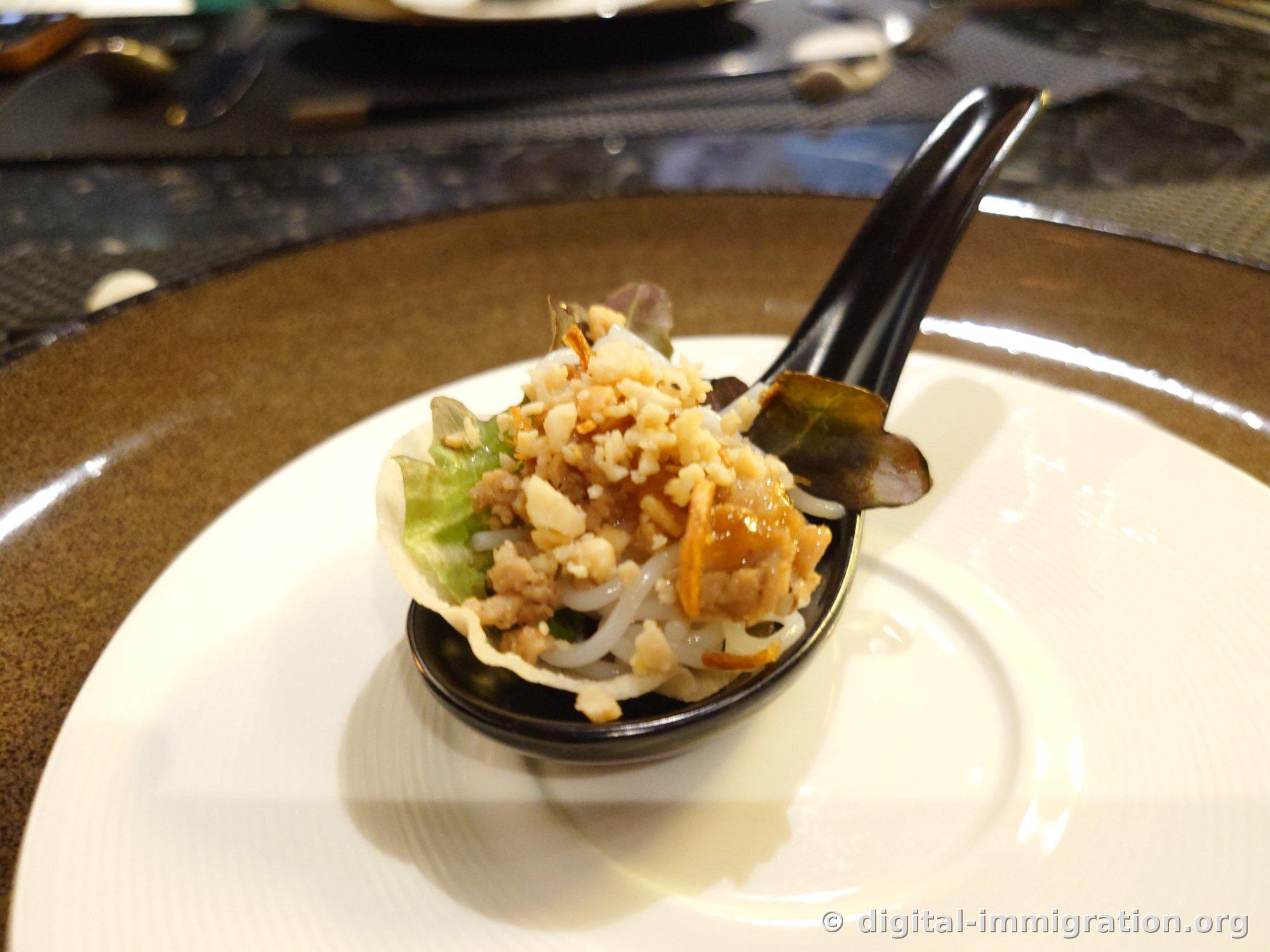 Single bite amuse-bouche on a black spoon topped with rice paper cup, minced pork, bean sprouts, and crushed peanuts