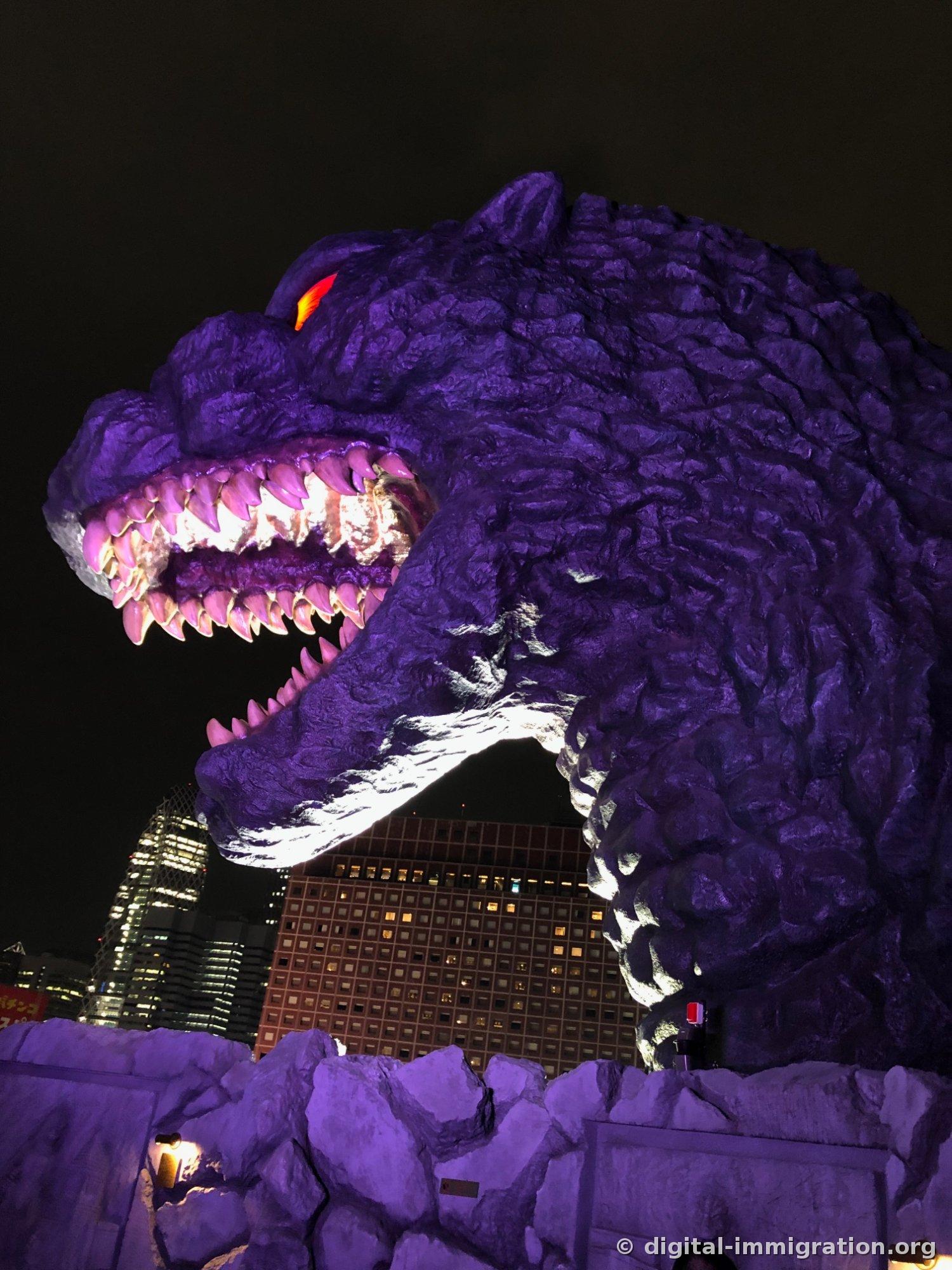 Gros plan de la tête de Godzilla illuminée en violet la nuit. La lumière brille dans les dents de la gueule ouverte, les yeux brillent en orange, et les gratte-ciel illuminés de Shinjuku sont visibles en arrière-plan