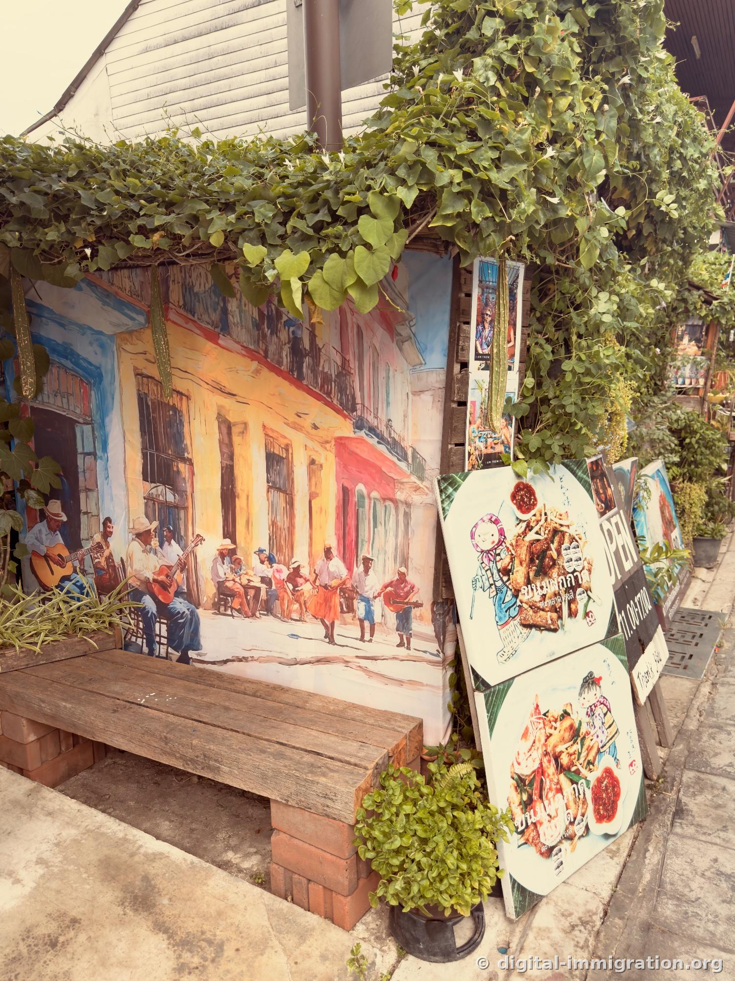 Wooden bench under vines with large painting of guitarist street musicians, Thai food menu board standing nearby