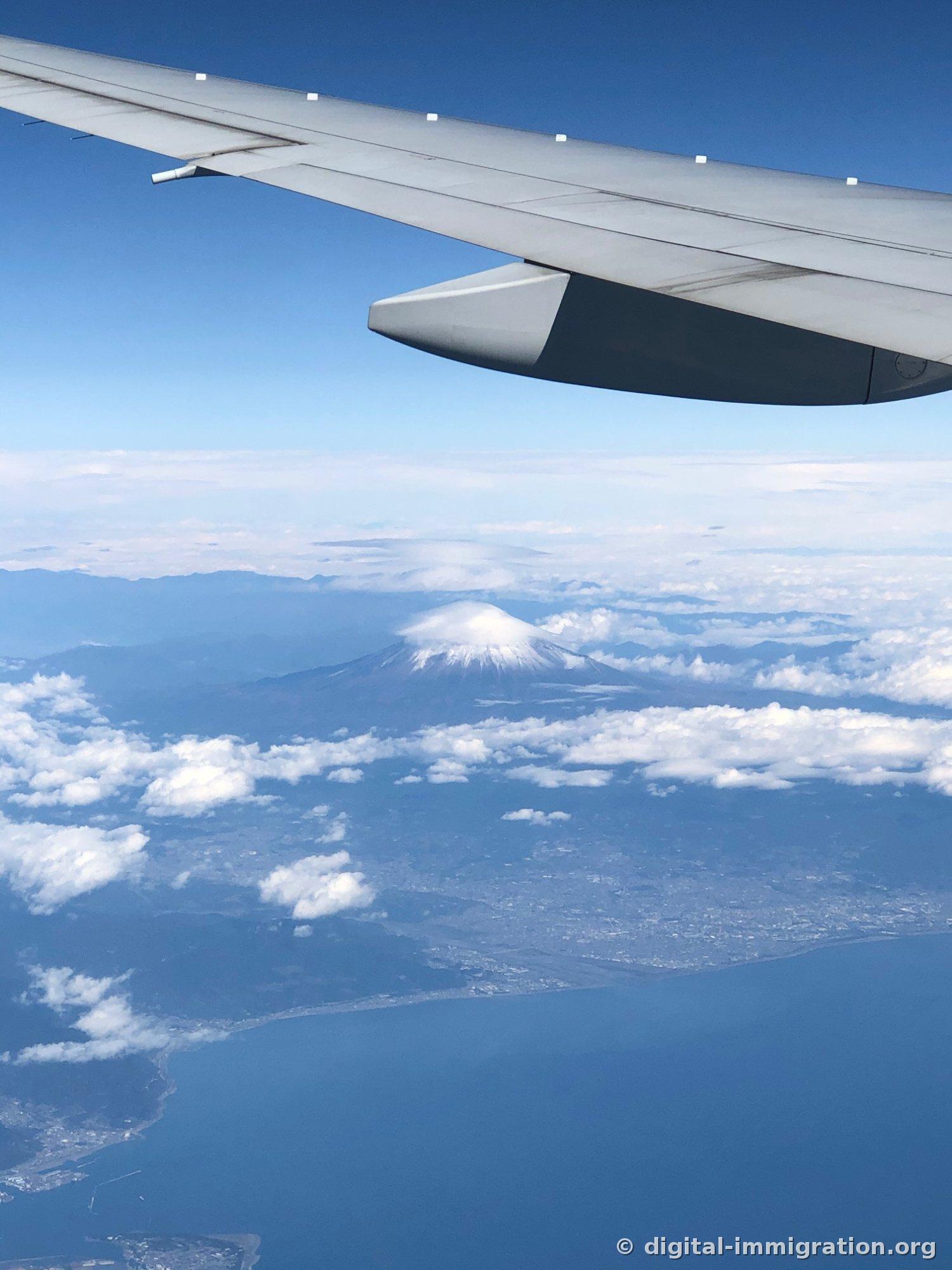 Une vue aérienne du Mont-Fuji visible sous l'aile de l'avion, avec un nuage blanc en forme de chapeau sur le sommet et les lignes côtières de la baie de Suruga et les villes qui s'étendent en dessous