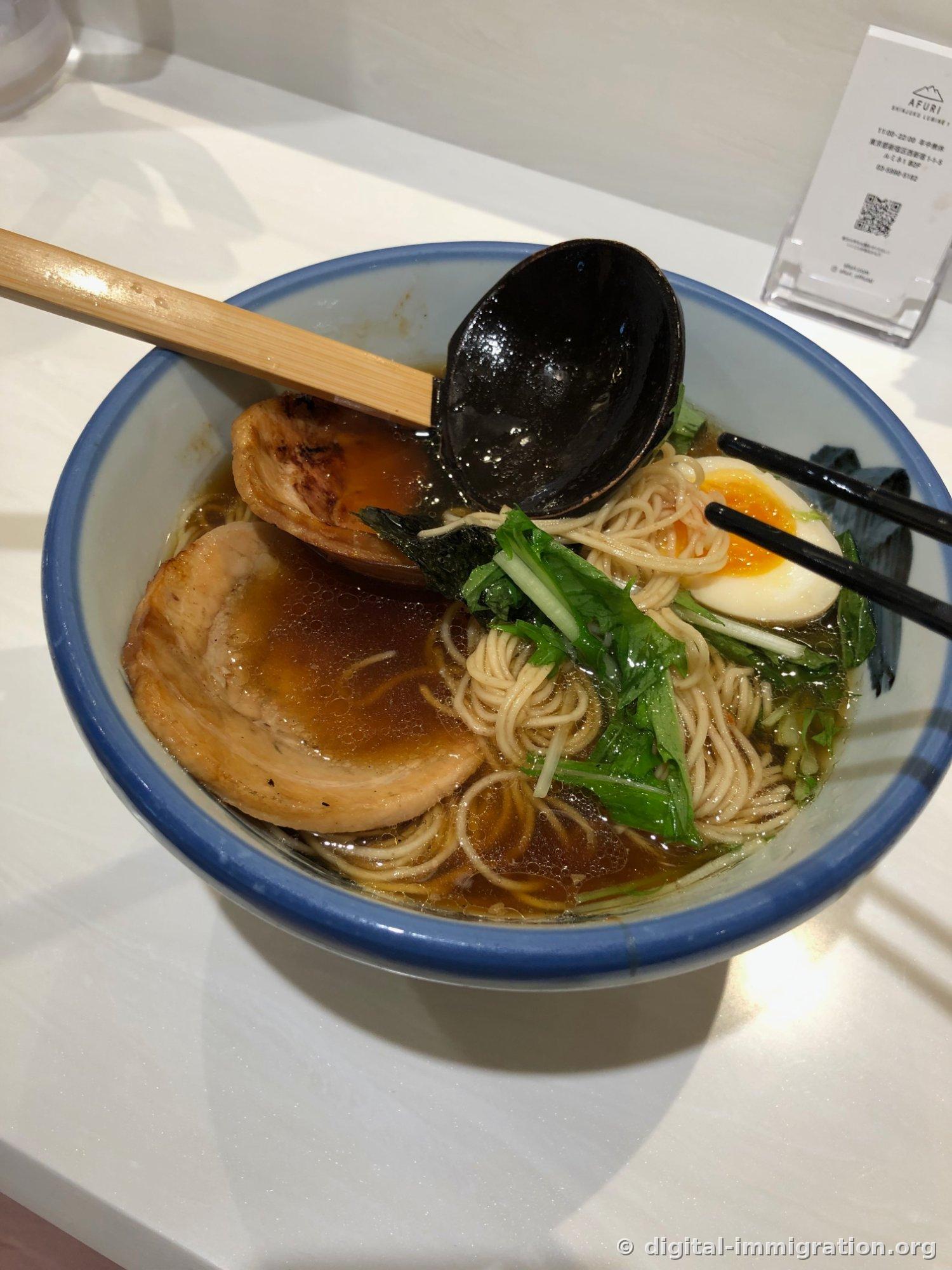 Ramen in a blue-rimmed bowl with clear broth, two pieces of torch-marked chashu, soft-boiled egg, mizuna vegetables, and a black ladle