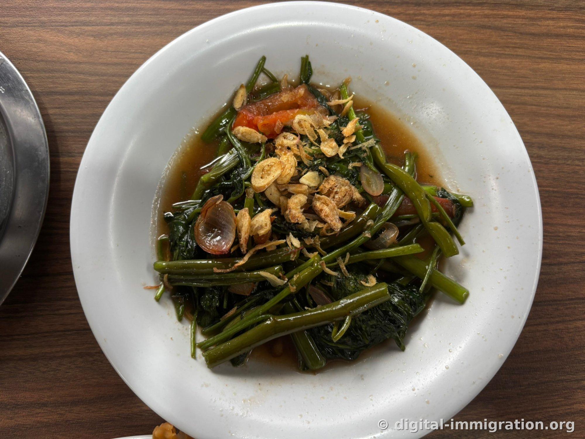 Water spinach stir-fry in soy broth on a white plate, with tomato pieces and fried shallots on top