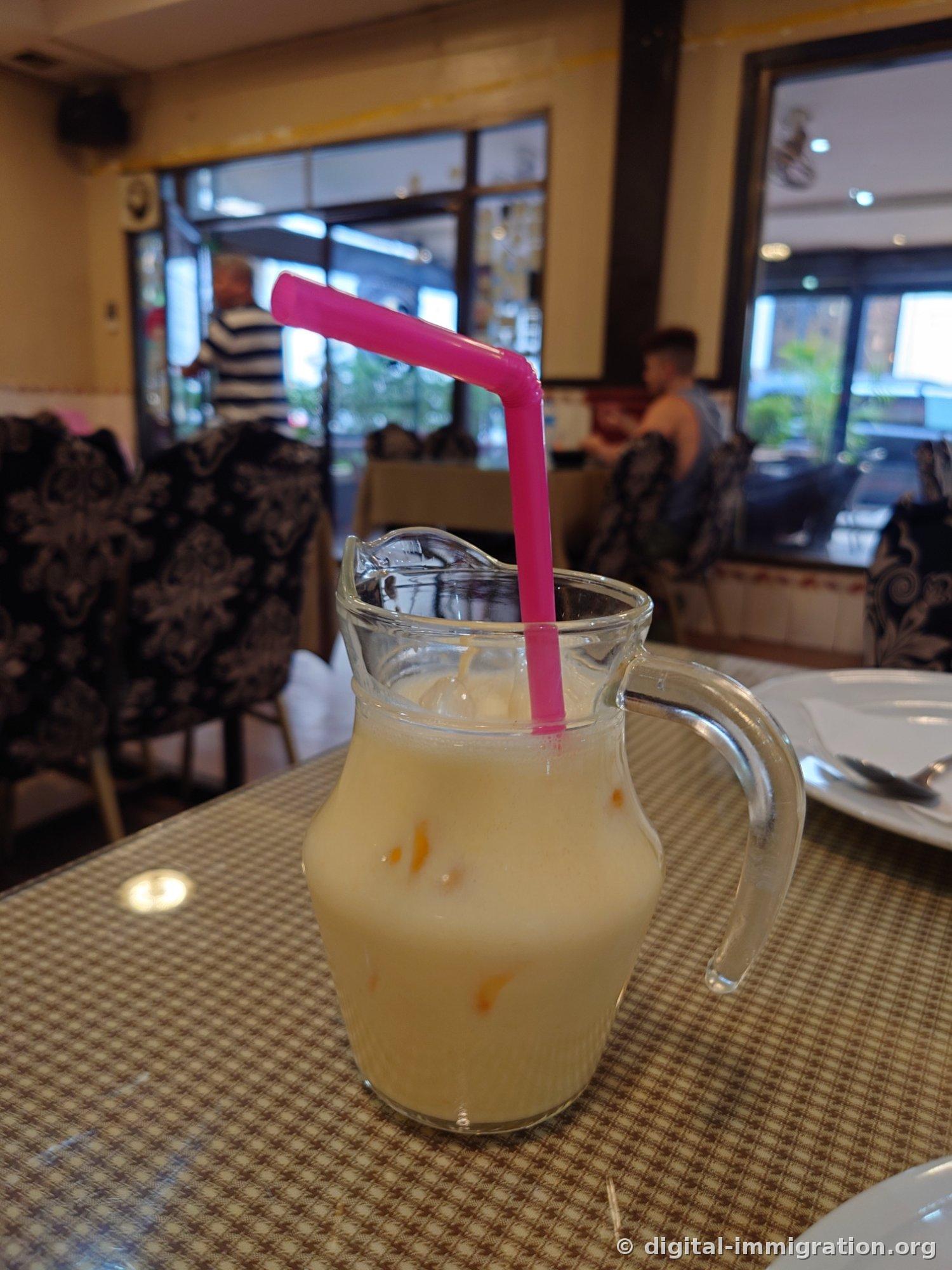 A glass pitcher filled with yellow mango lassi and a pink straw, with blurred restaurant patrons visible in the background