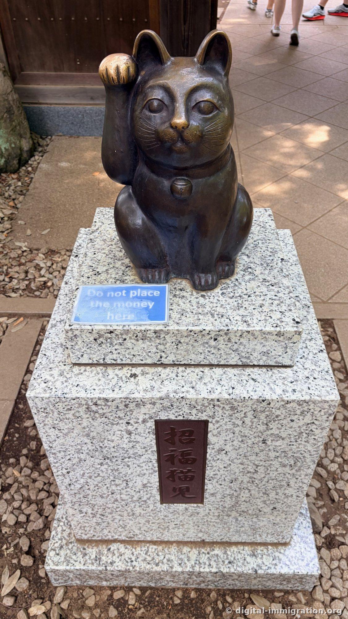 A bronze beckoning cat statue on a granite base, with a nameplate reading 'Shofuku Byoa' and an English notice saying 'Do not place the money here'