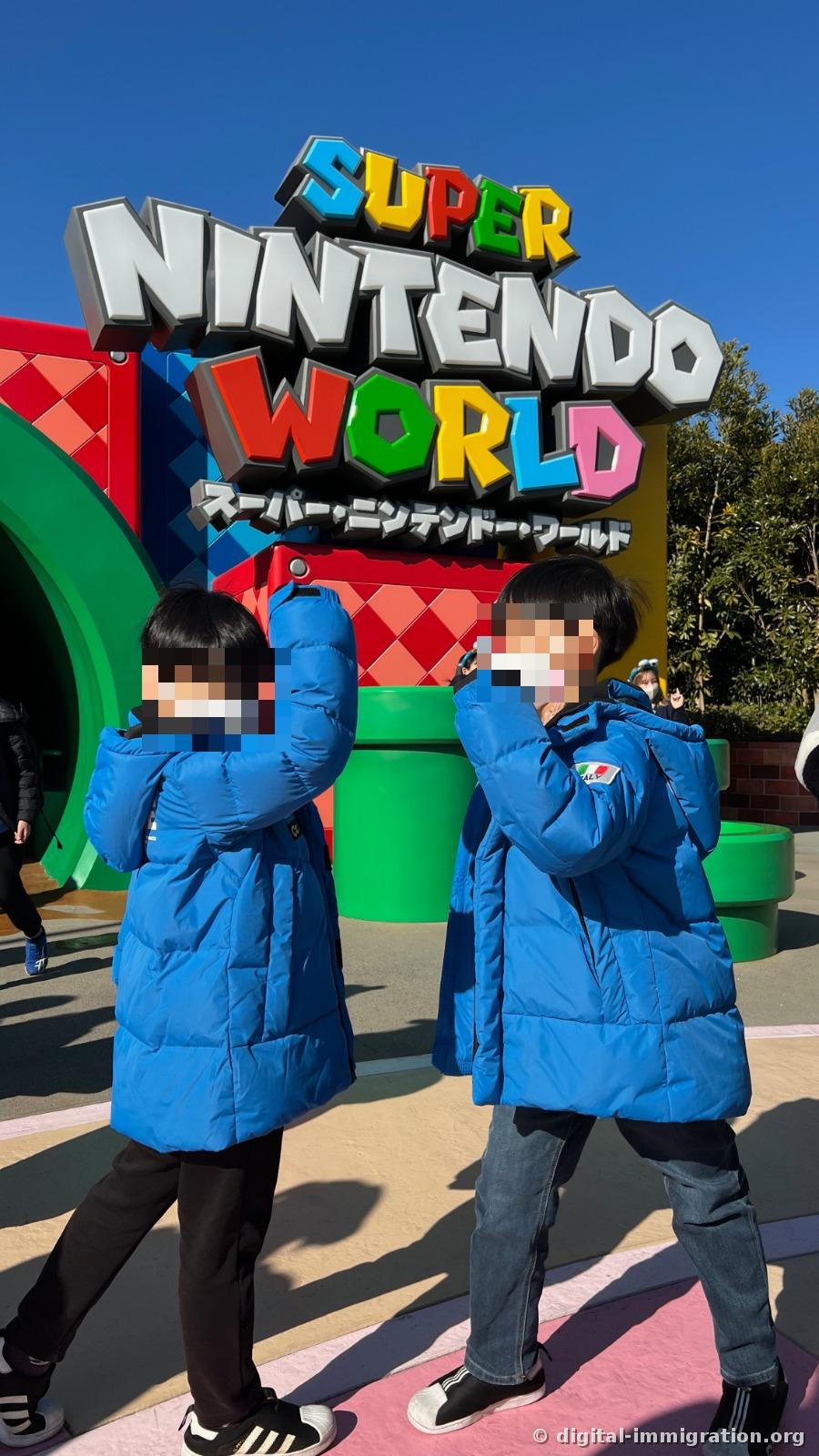 Two children in blue padded jackets are posing as if jumping beneath the large 'SUPER NINTENDO WORLD' sign in front of the green pipe sculpture