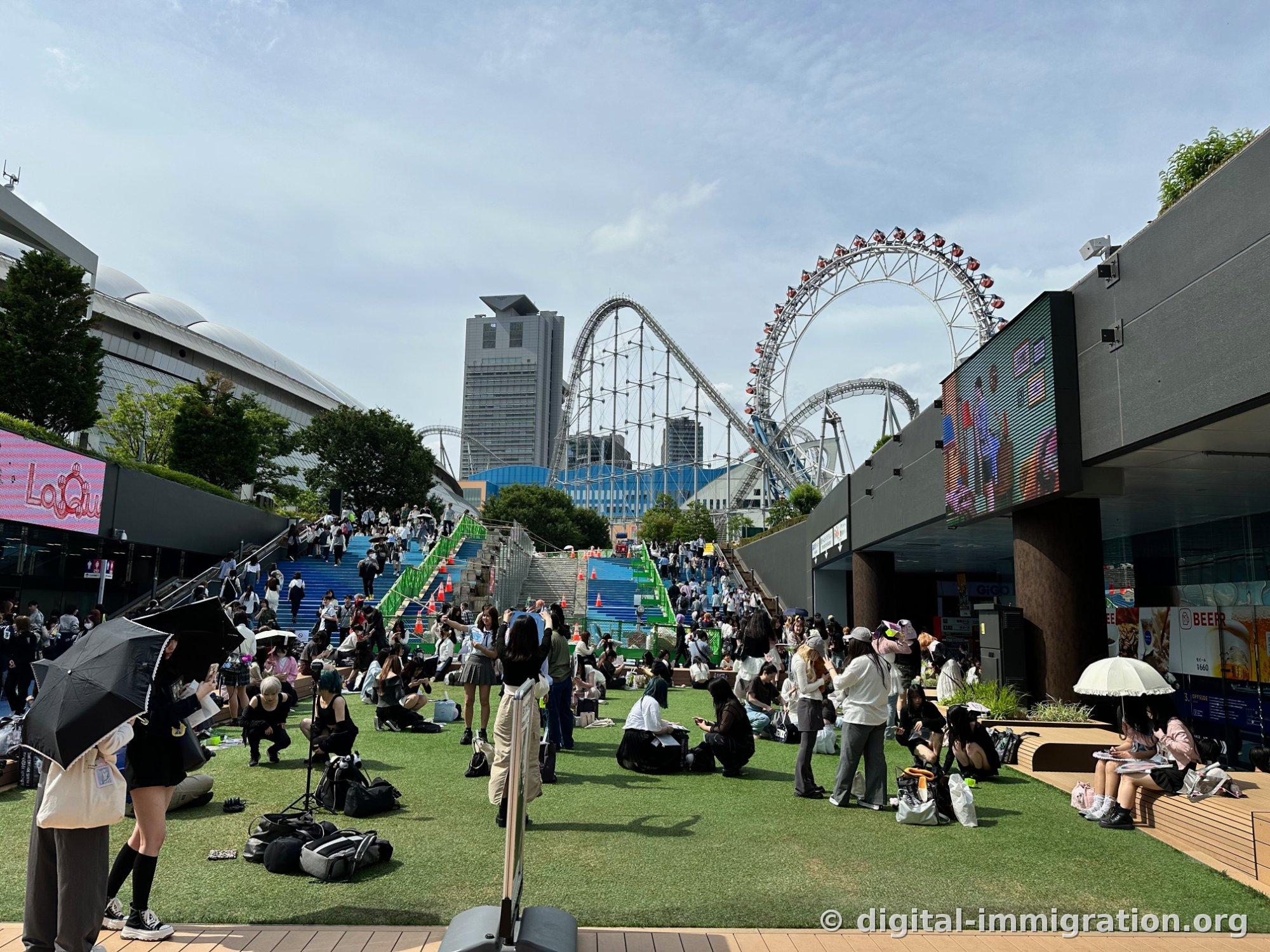 Des gens se rassemblent sur la pelouse artificielle, et à l'arrière on voit l'escalier bleu et la pente verte, au-delà le toit blanc du dôme de Tokyo et les rails du manège à sensations Thunder Dolphin, ainsi que la grande roue Big O