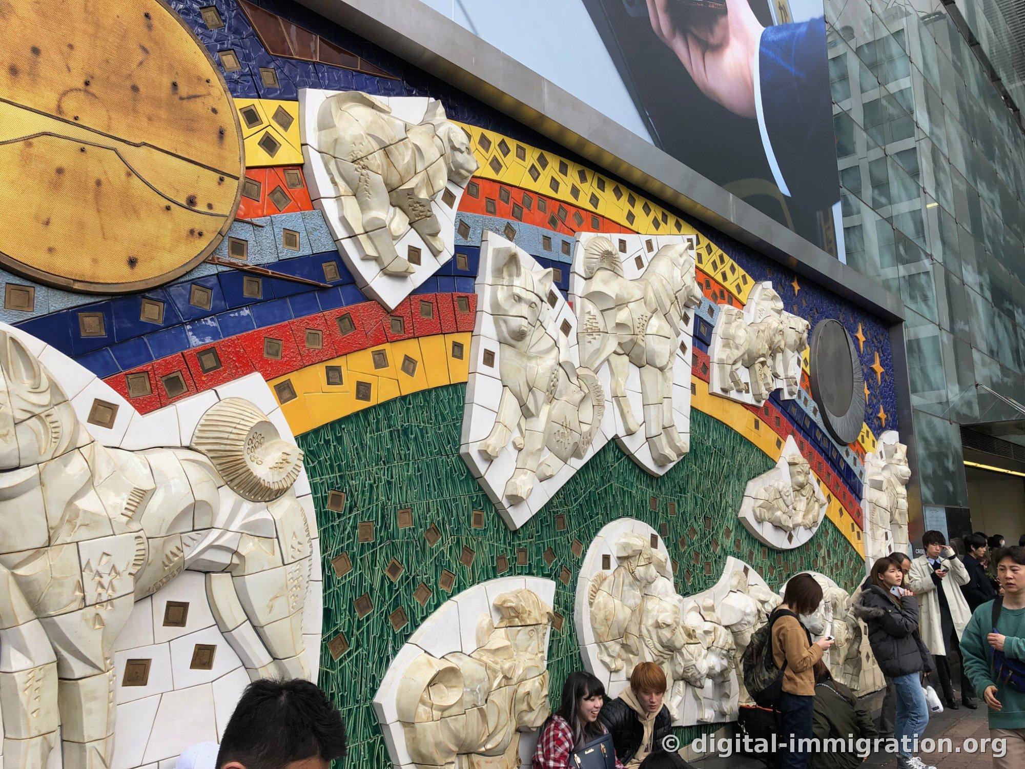 虹色のタイルの上に白色の犬の浮き彫りが複数付いている渋谷駅の大型壁画、下に人々が座っている