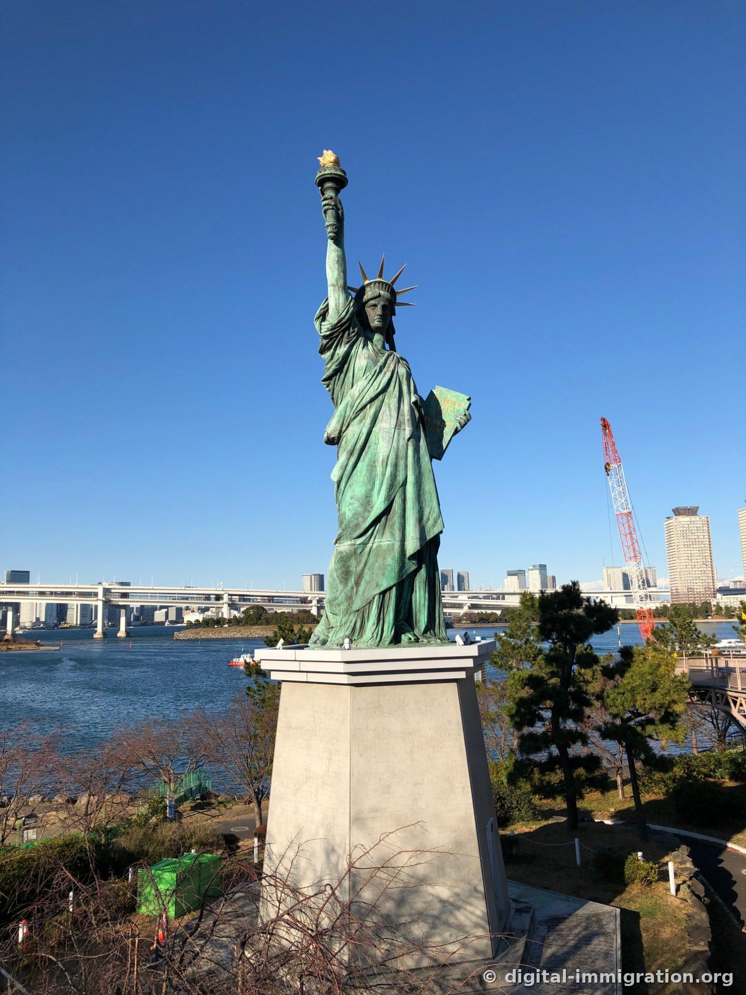 蓝天下青铜生锈的台场自由女神像正面，背景是东京湾海面和彩虹大桥桥面，右侧可见红色施工吊车
