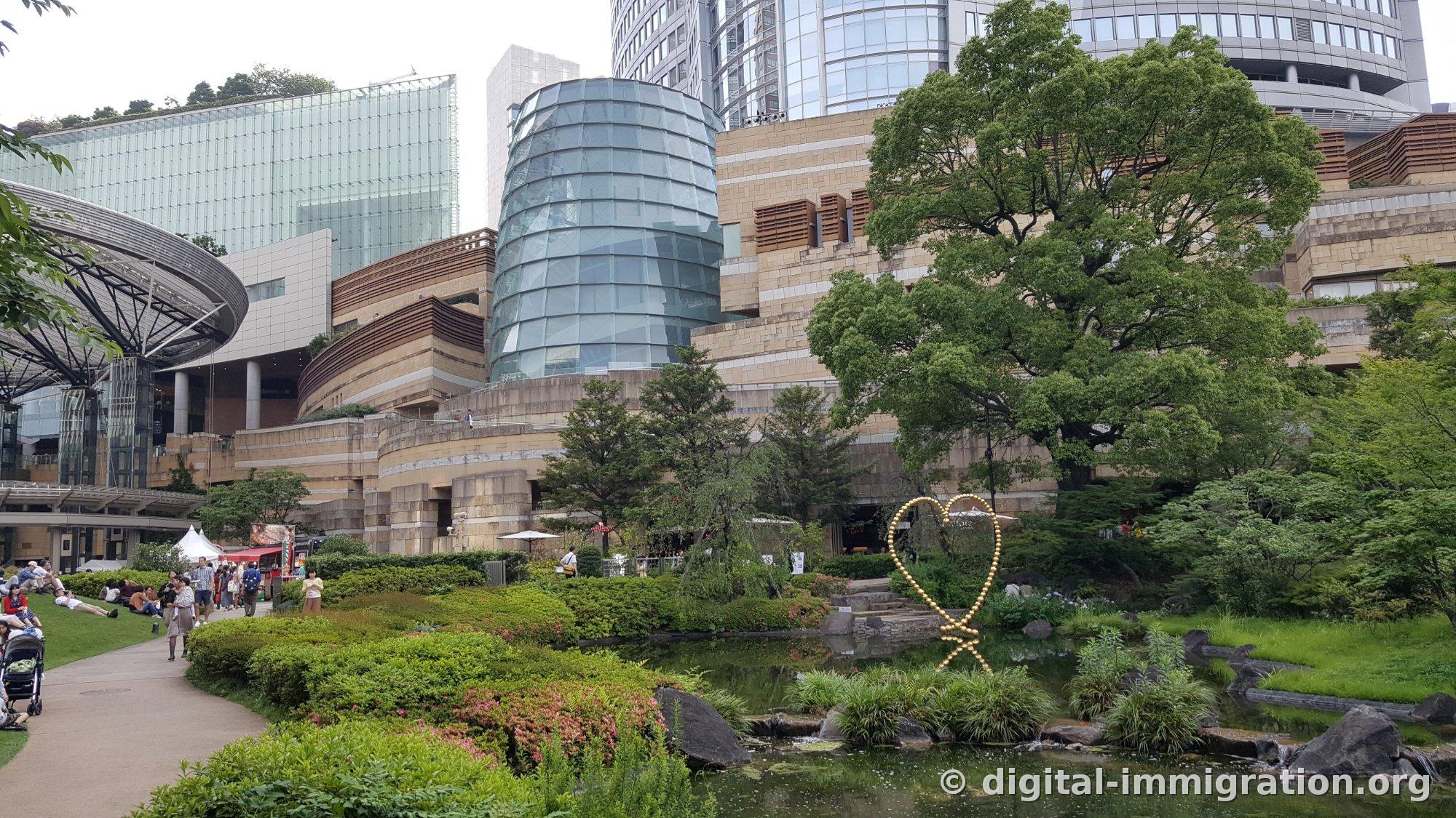 L'étang et les arbres verts du jardin Mori de Roppongi Hills, avec un bâtiment cylindrique en verre et une haute tour se chevauchant à l'arrière, et une sculpture en forme de cœur doré visible près de l'étang à droite