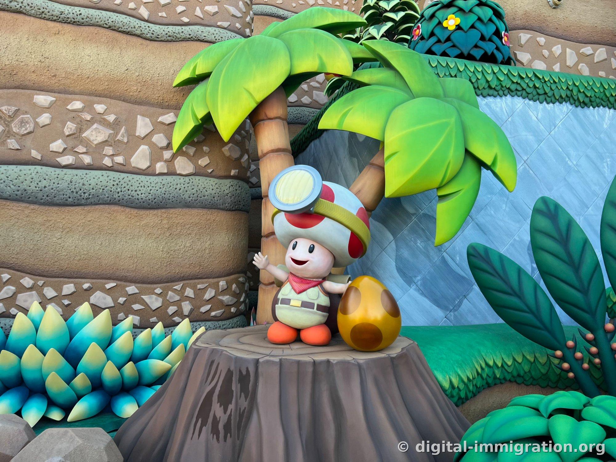 Captain Toad sculpture with raised hand standing on a tree stump beneath a palm tree, and a brown spotted egg beside it