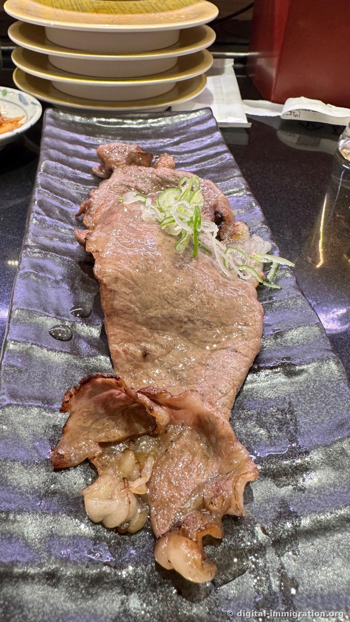 深蓝色长方形盘子里铺着薄片牛肉烤和葱段，后面堆放着吃完的四张黄色盘子