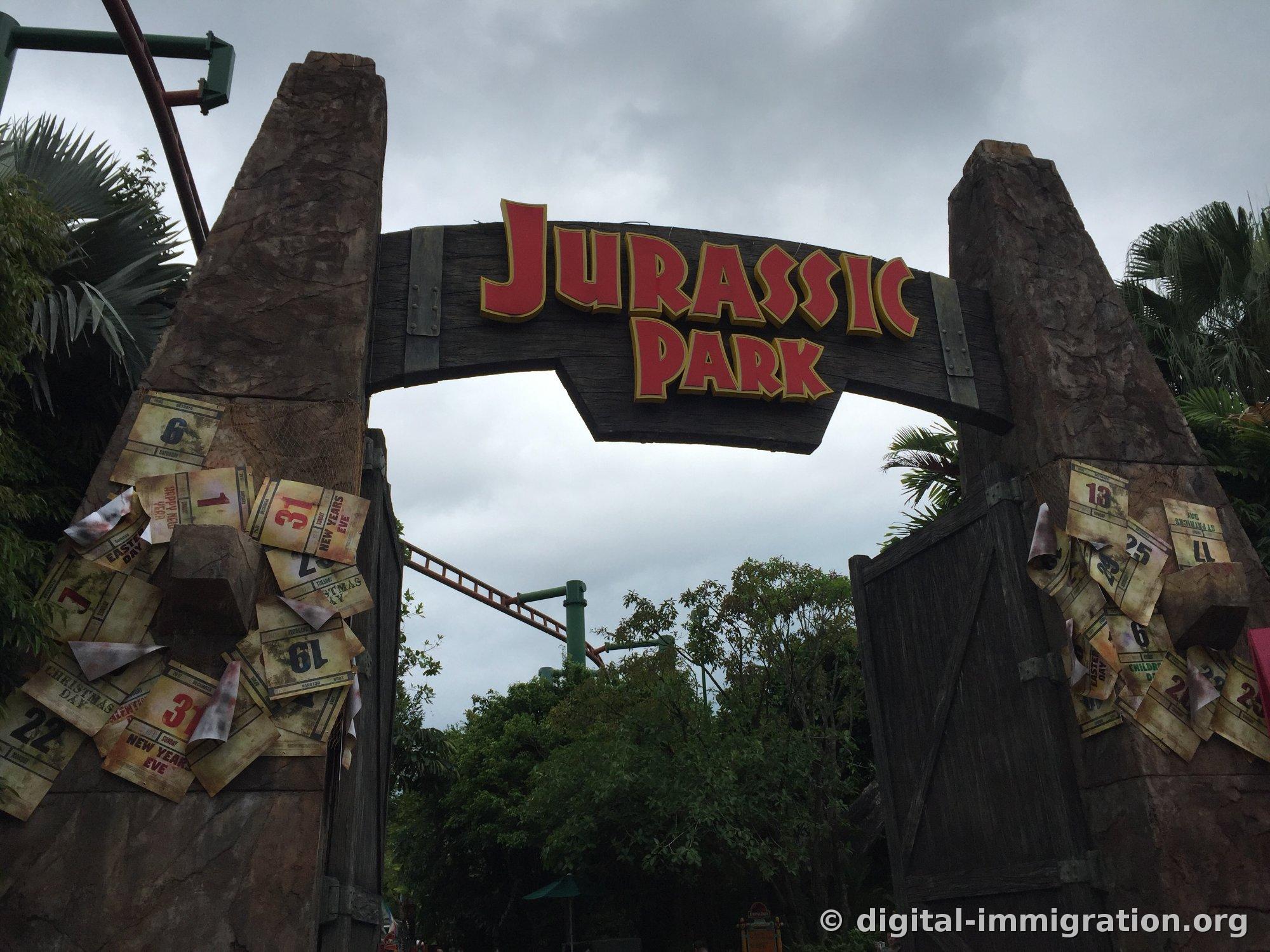 Jurassic Park gate made with trees and rocks under an overcast sky, with a red JURASSIC PARK sign and torn calendar paper decoration