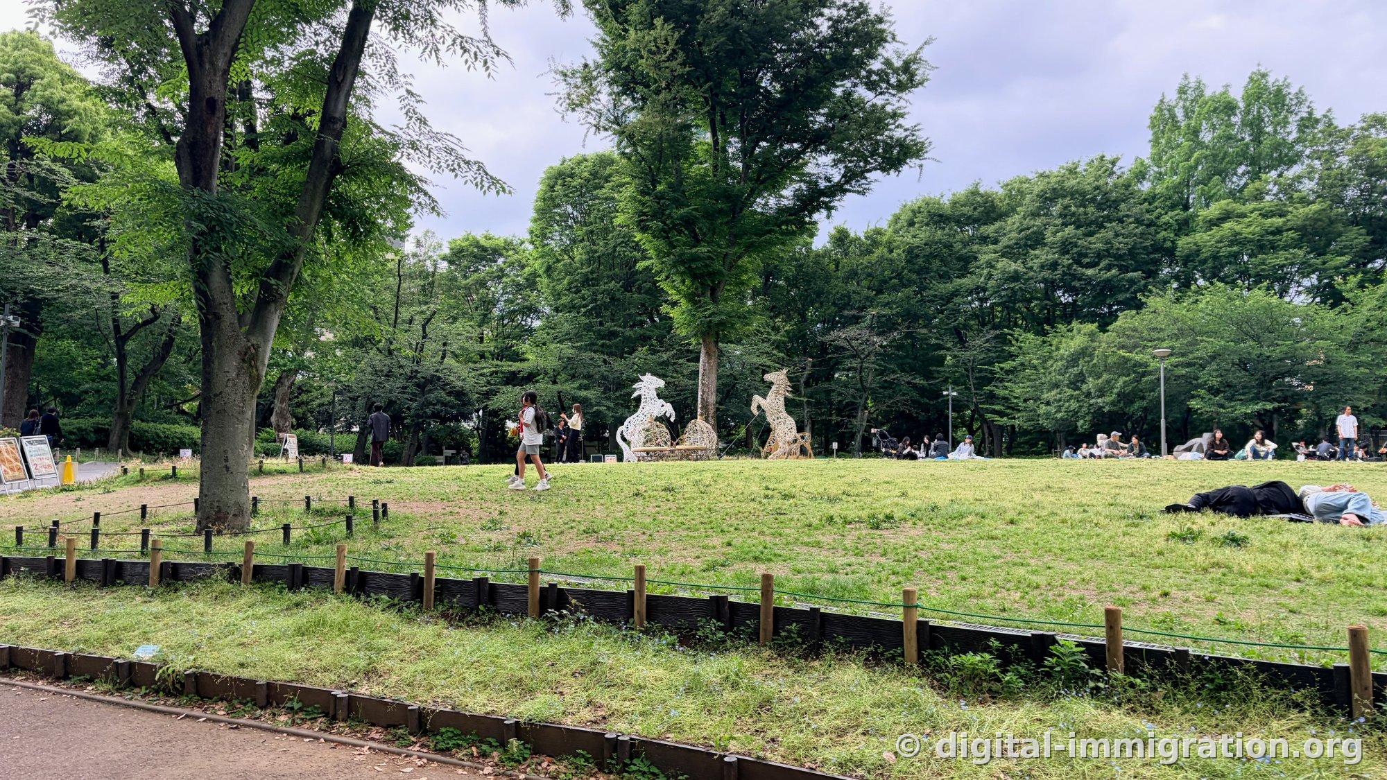 Deux sculptures de chevaux blancs debout sur les pattes arrière sur une large pelouse avec des gens assis ou allongés tout autour, séparés par des piquets de bois bas et des cordes vertes