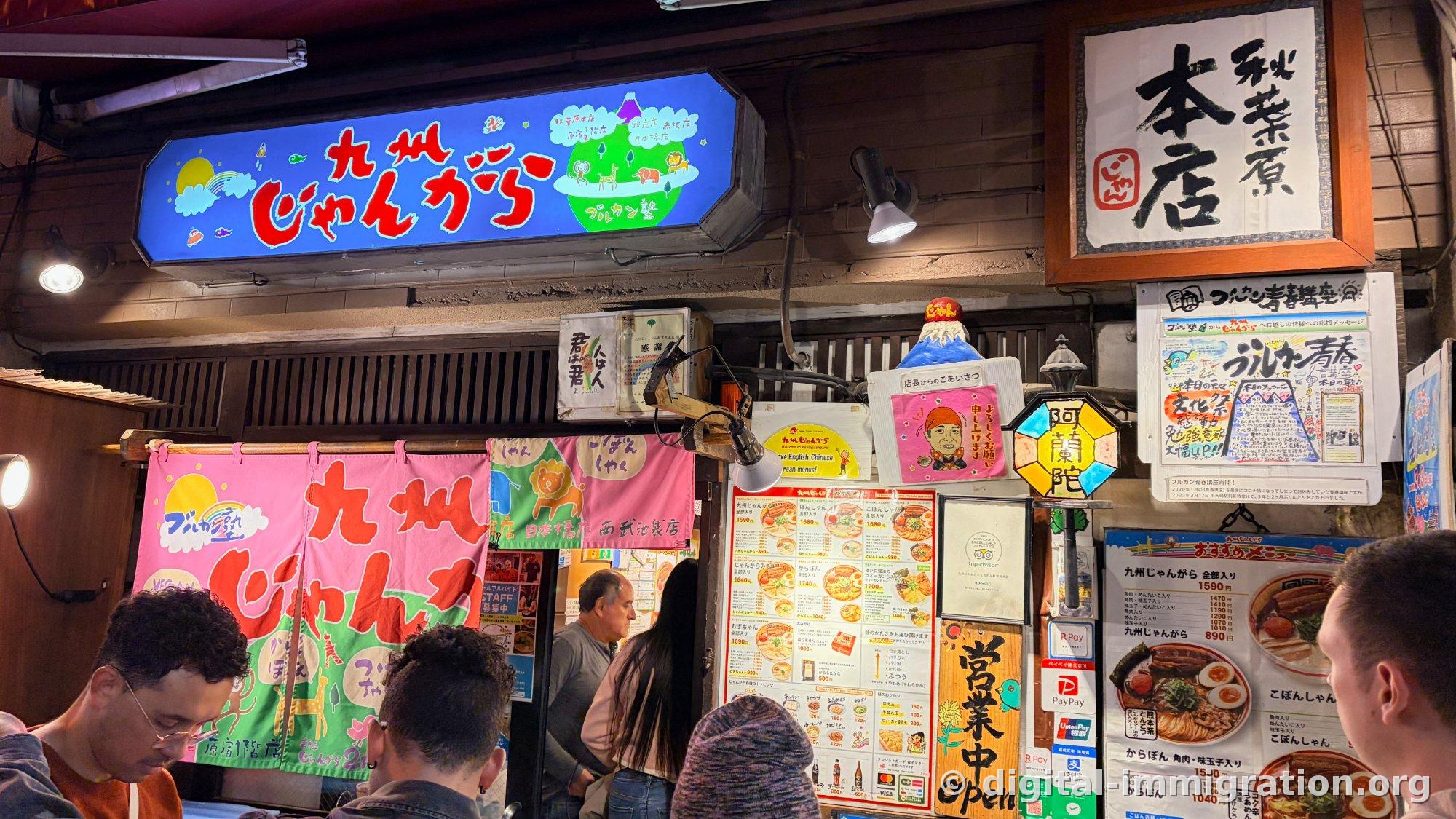 The entrance to the Akihabara main branch with a signboard reading Kyushu Jangara in red letters on a blue background, a framed brush-stroke calligraphy reading Akihabara Main Branch, a pink noren curtain, and customers lined up
