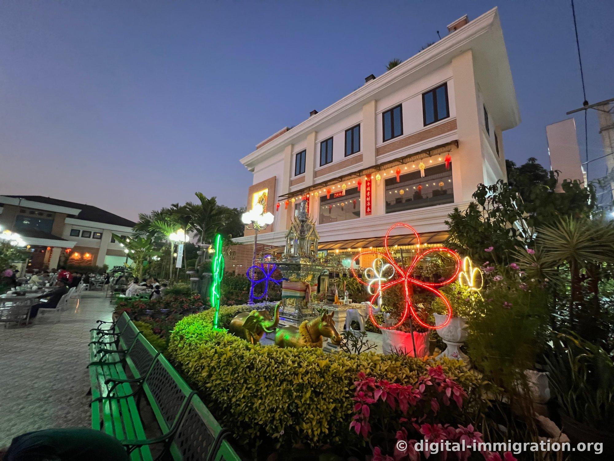 A spirit house with golden elephant and horse sculptures in the courtyard flower bed, surrounded by red, blue, and green neon flower decorations, with a two-story building hung with red lanterns in the back