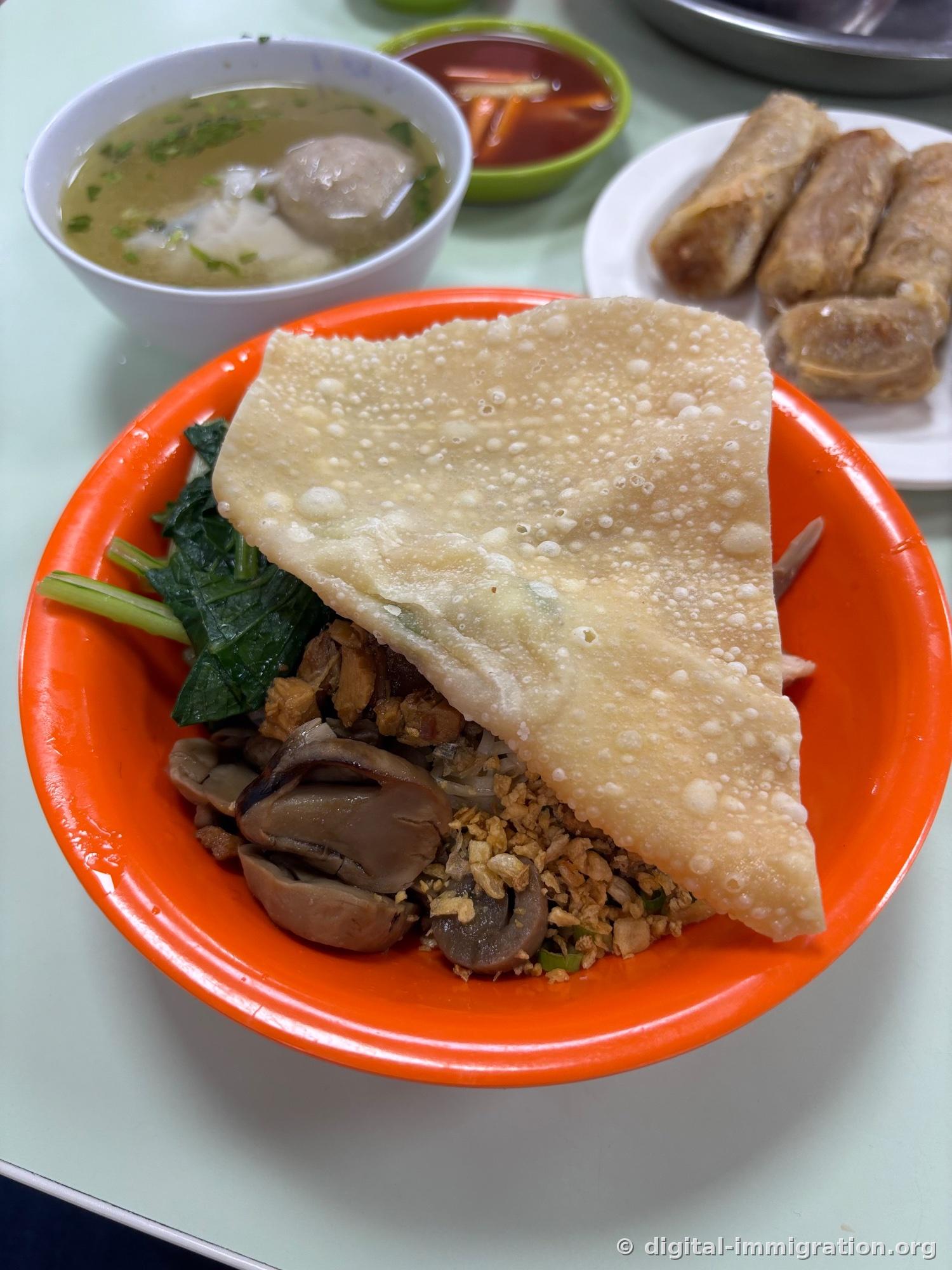 Bol orange de bakmi couvert d'une feuille de pâte frite de la taille d'une paume, avec champignons shiitake, bok choy et ail frit, bouillon de wonton et assiette de fritures en arrière-plan