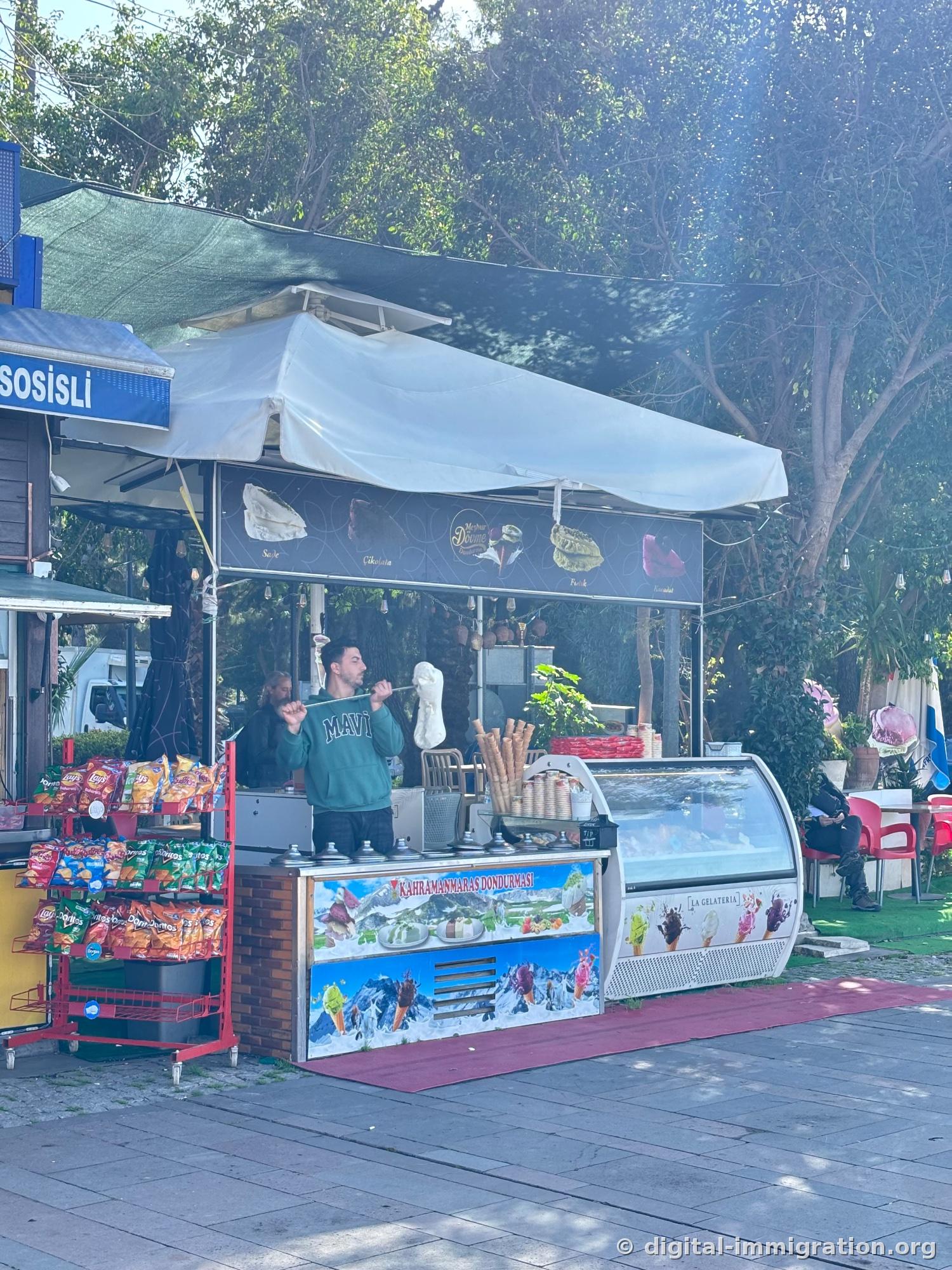 Vendor lifting stretchy Kahramanmaraş Dondurması ice cream on a long metal rod at harbor-side stall, snack display and ice cream showcase beside