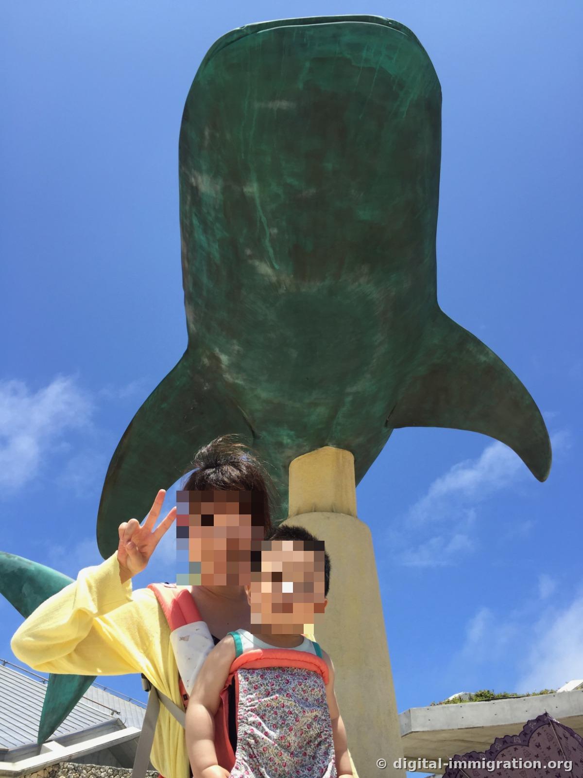 从下往上拍的青铜鲸鲨雕像腹部和尾鳍，一个做V字手势的人和一个婴儿站在前面