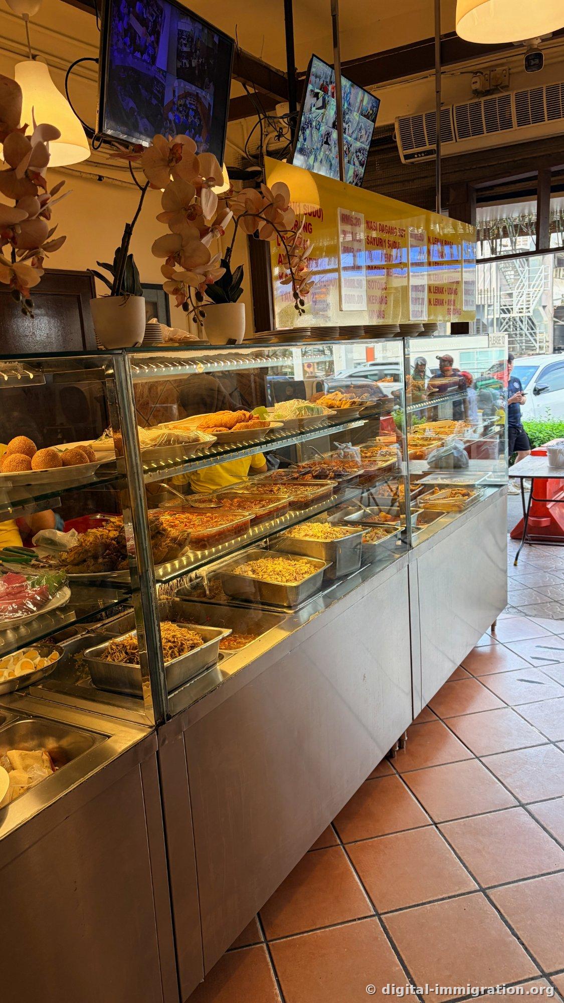 Two CCTV monitors hang from the ceiling, and below them a price board with red text on a yellow background listing menu items and prices, and below that a long illuminated glass display case extends into the shop