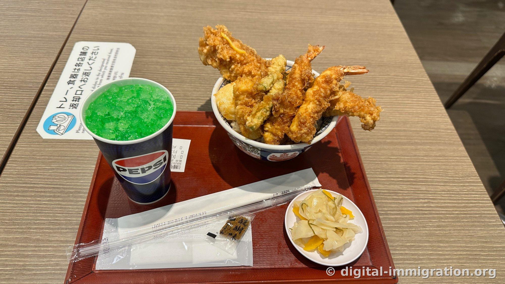 A fried shrimp donburi set on a red tray with a fluorescent green Pepsi melon soda paper cup, a plate of pickled vegetables with yellow pickled radish, chopsticks and seasoning packets, a tray return instruction card visible in back