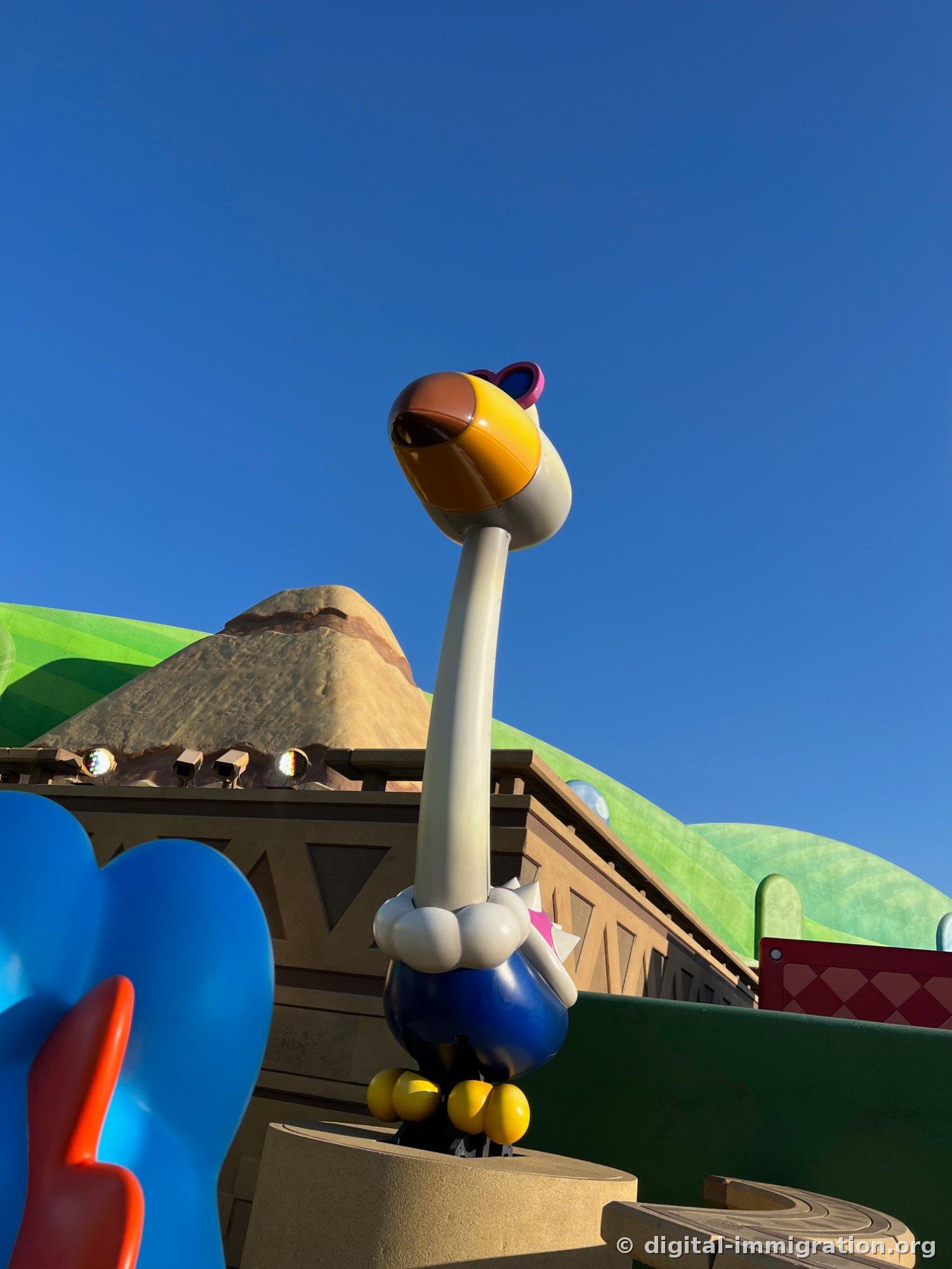 A long-necked bird-shaped sculpture with a yellow beak and goggles stands in front of a desert-style building against a blue sky