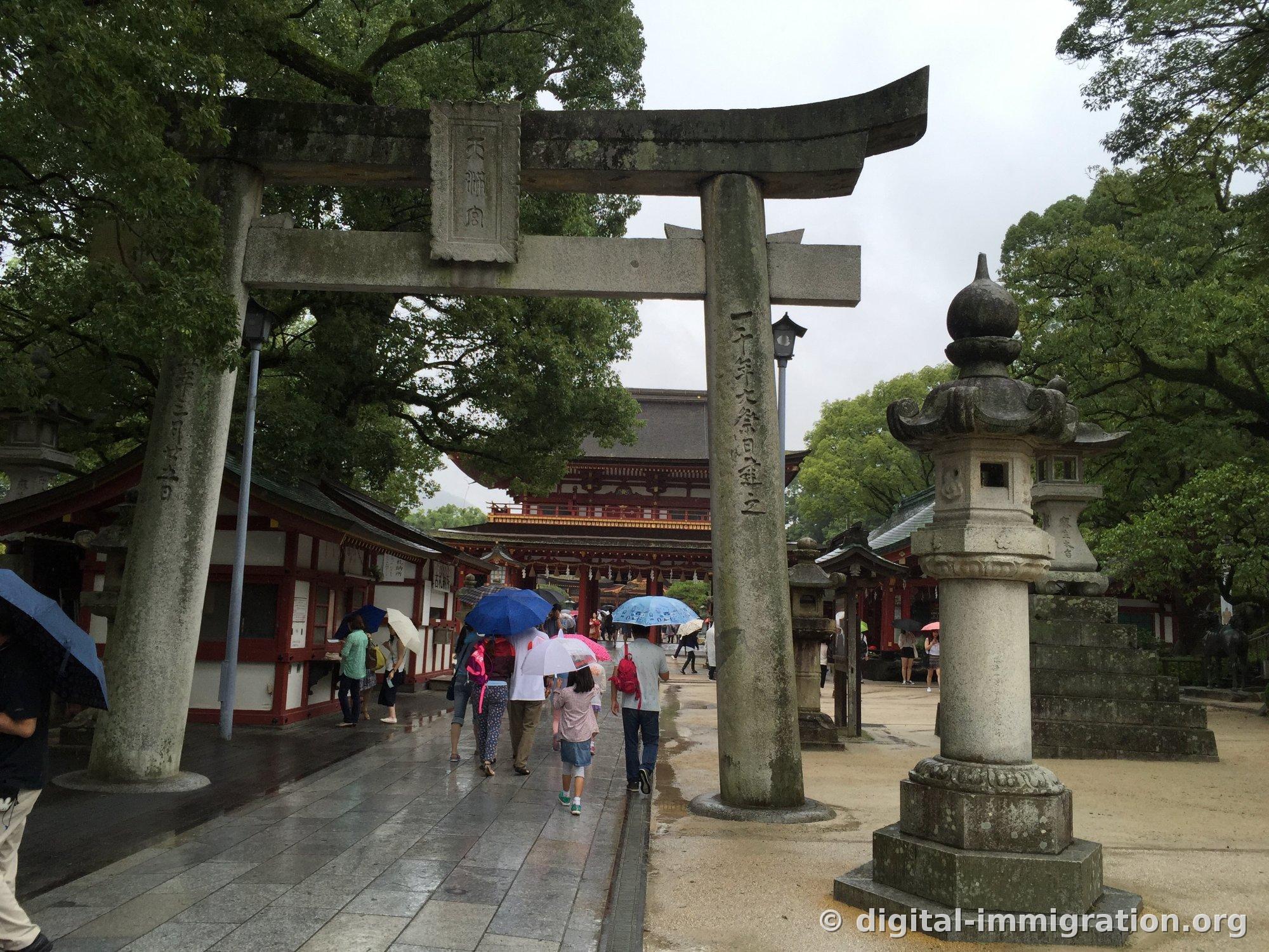 Beyond a stone torii with the inscription '天満宮', a red romon gate is visible, along with a stone lantern on the approach path