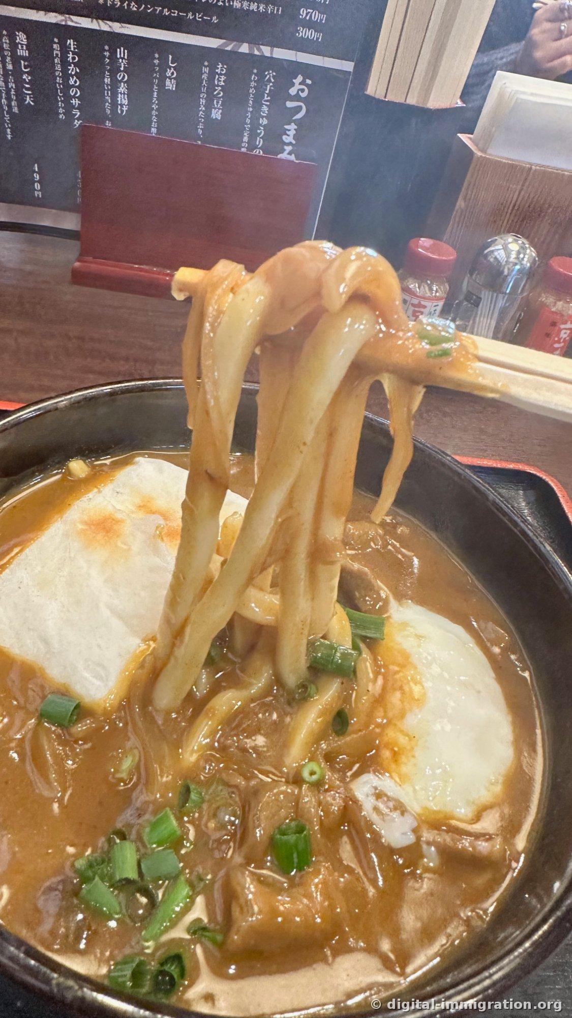 Les nouilles udon épaisses soulevées avec les baguettes, avec un bouillon curry épais qui colle, et en bas le bol avec l'aburaage et l'œuf. À l'arrière, le menu d'accompagnements noir et les condiments