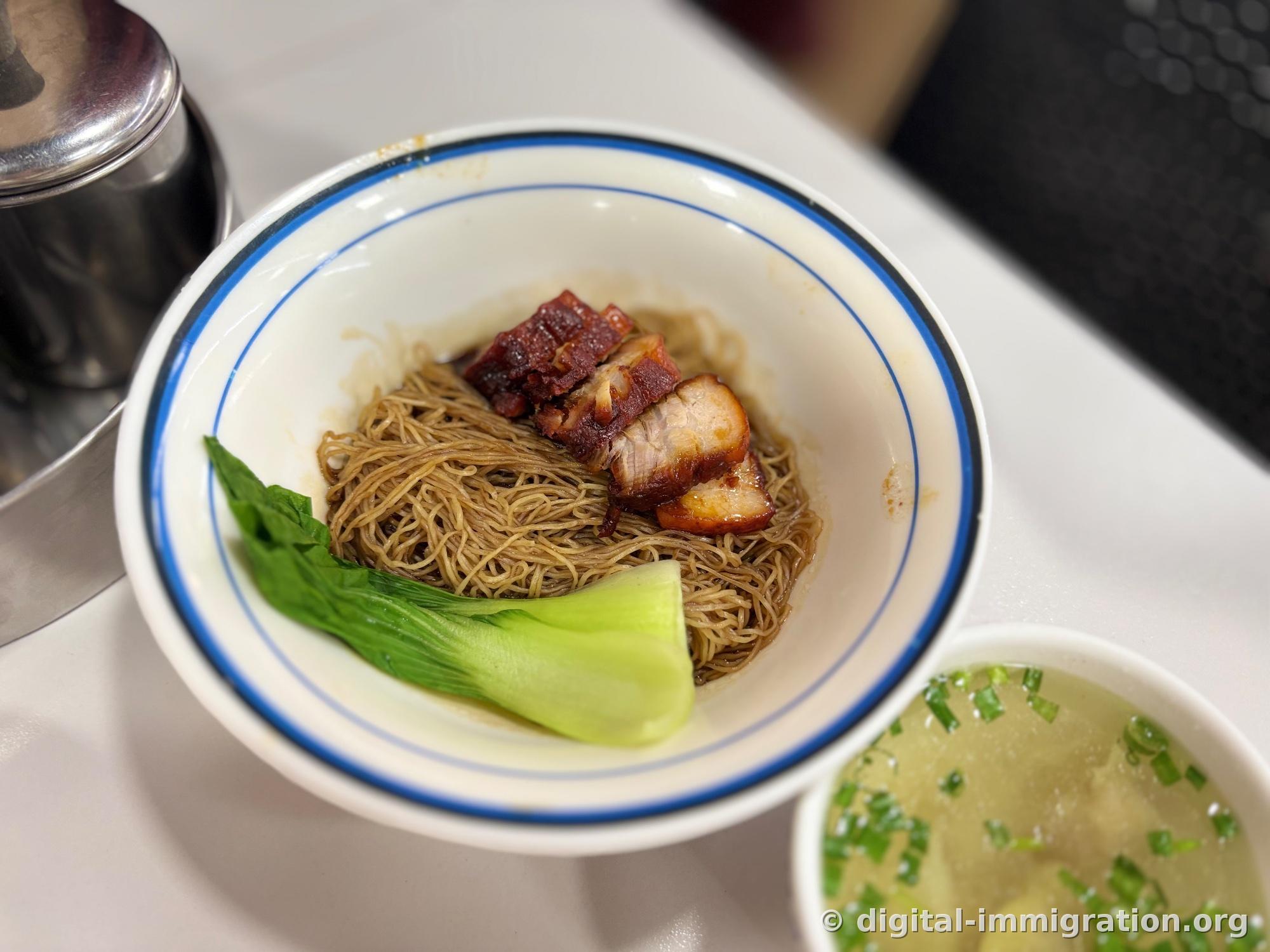 Bowl with blue rim holding dry wanton noodles. Thin noodles tossed in soy sauce topped with three pieces of char siu and blanched Chinese broccoli
