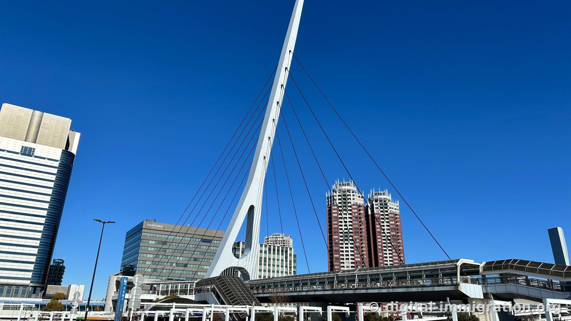A white cable-stayed bridge with black cables stretching across blue sky, a glass-roofed pedestrian passage below it, a building with SUNTORY signage, and a reddish-brown high-rise apartment building on the right