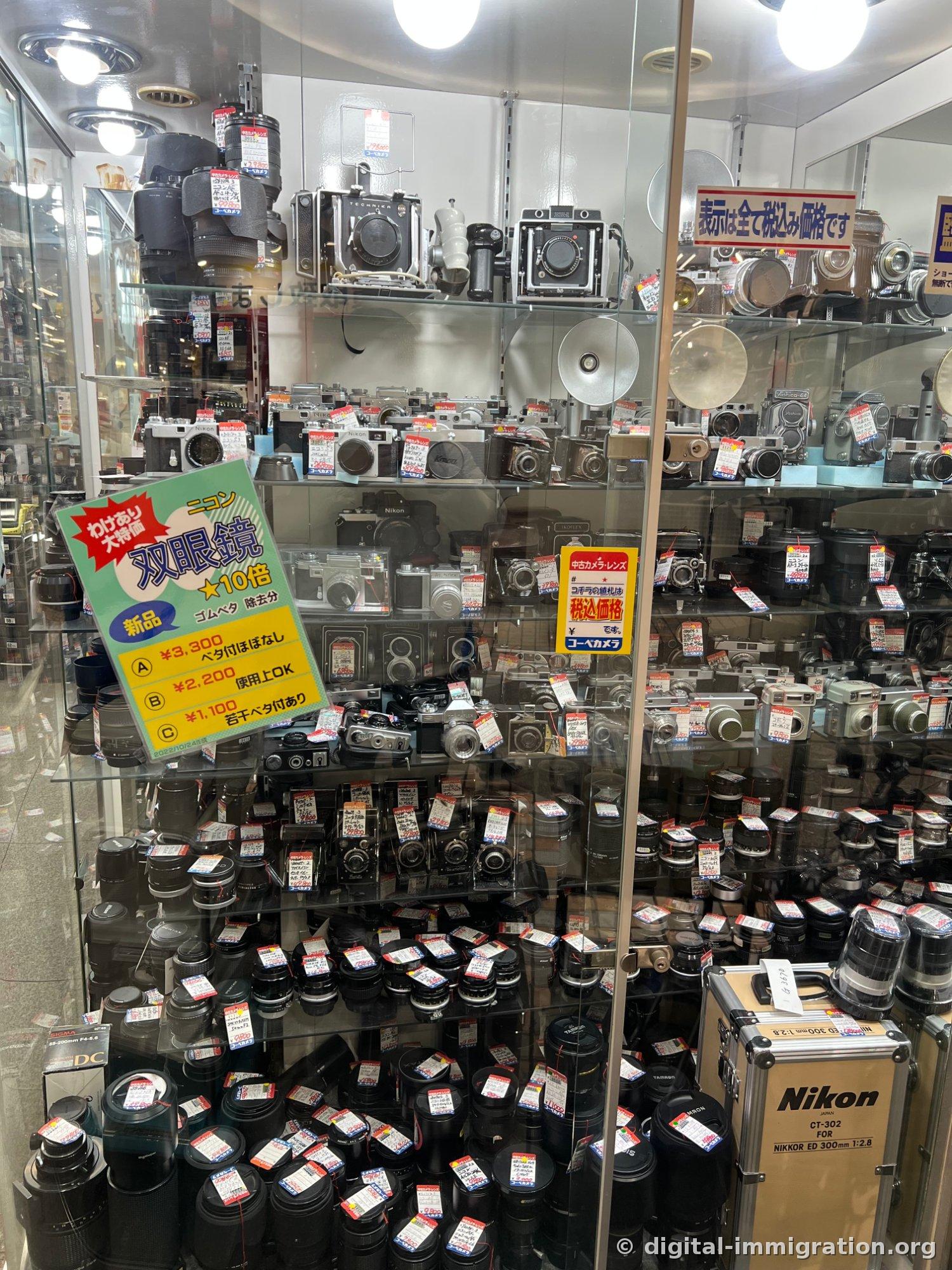 Une vitrine de magasin de caméras d'occasion en verre avec des boîtiers de caméras argentiques et des objectifs densément exposés en couches, chacun avec une étiquette de prix