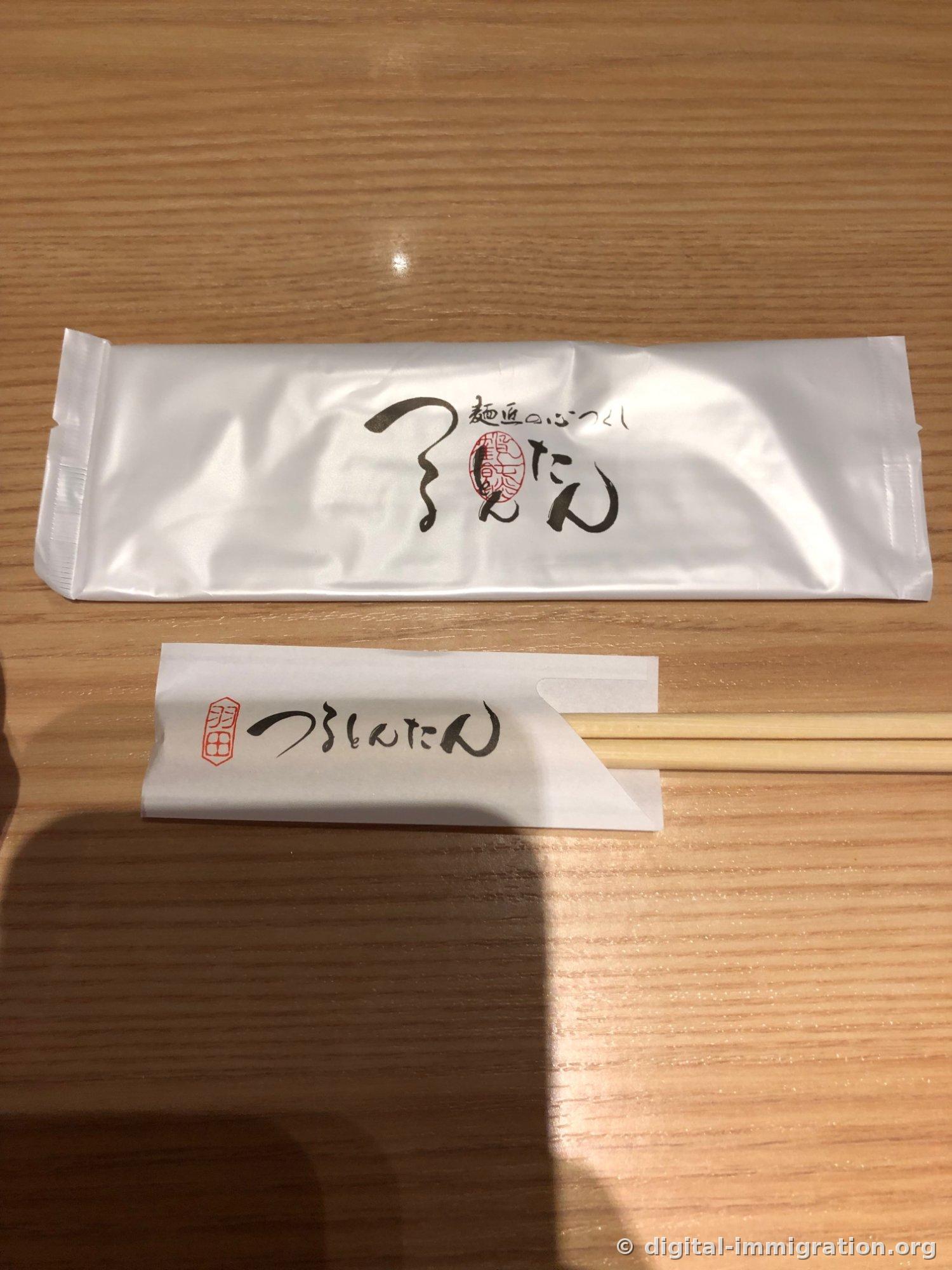 A white wet towel envelope with the Tsurutontan logo and a chopstick envelope stamped with red seal are placed on a wooden table
