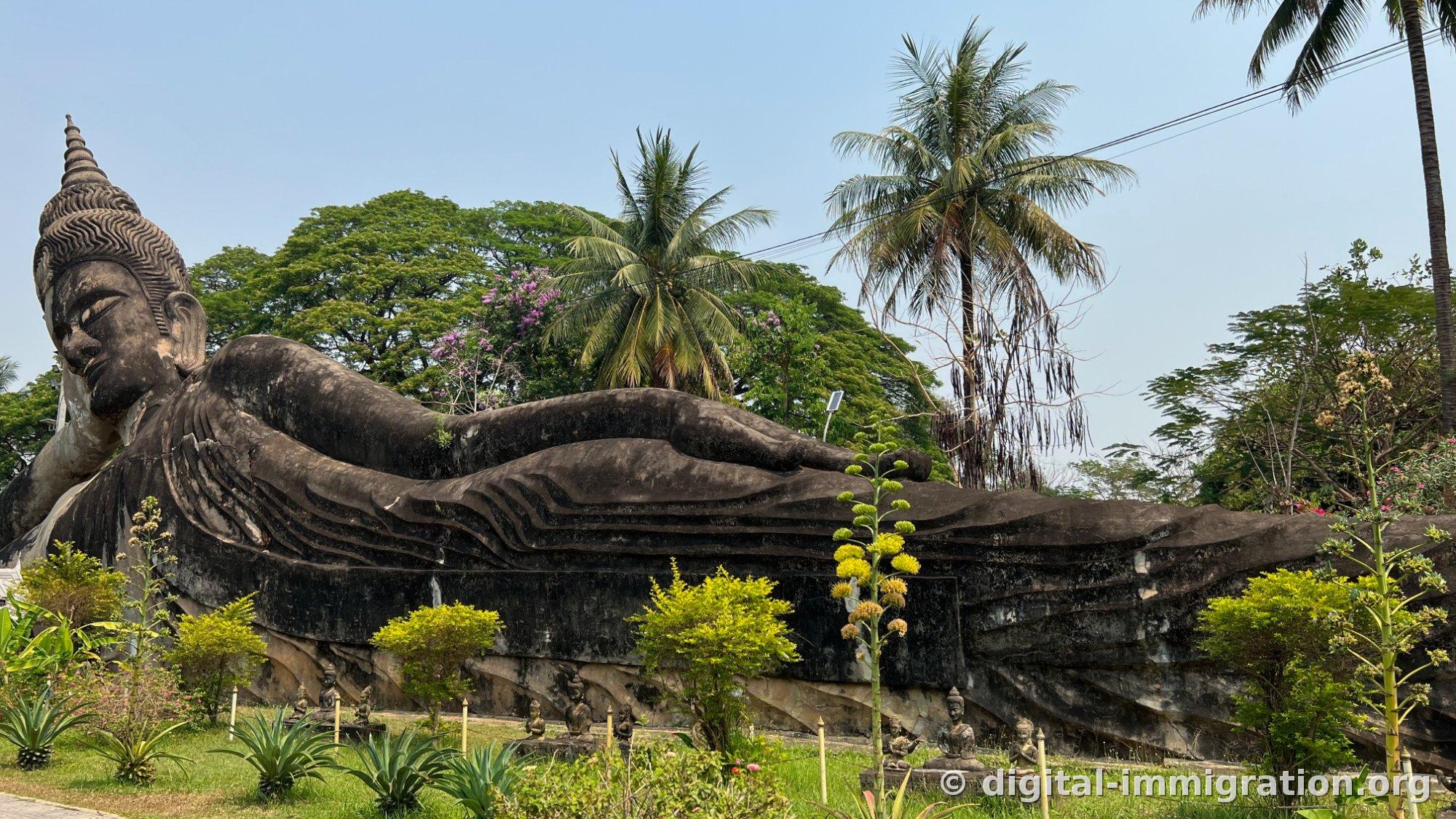 Un énorme Bouddha couché gris allongé sous les palmiers avec de petites statues alignées le long du socle