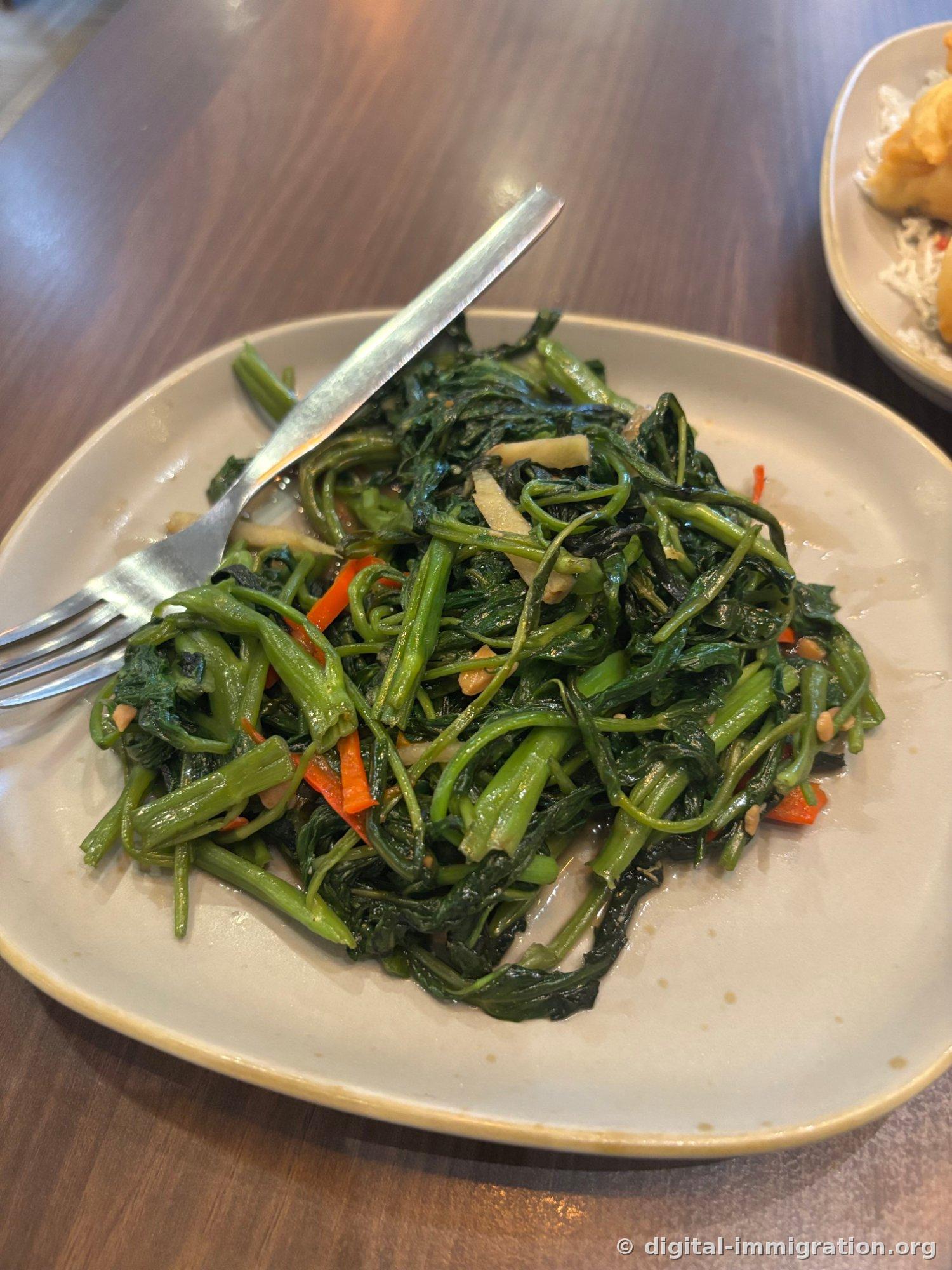 Morning glory stir-fried with sliced garlic and red chili, piled high on a plate with a fork resting on top