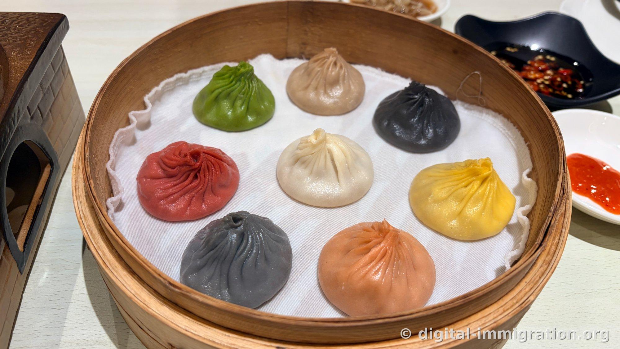 Eight-colored xiaolongbao in bamboo steamer basket with chili soy sauce and red chili sauce dishes on the side