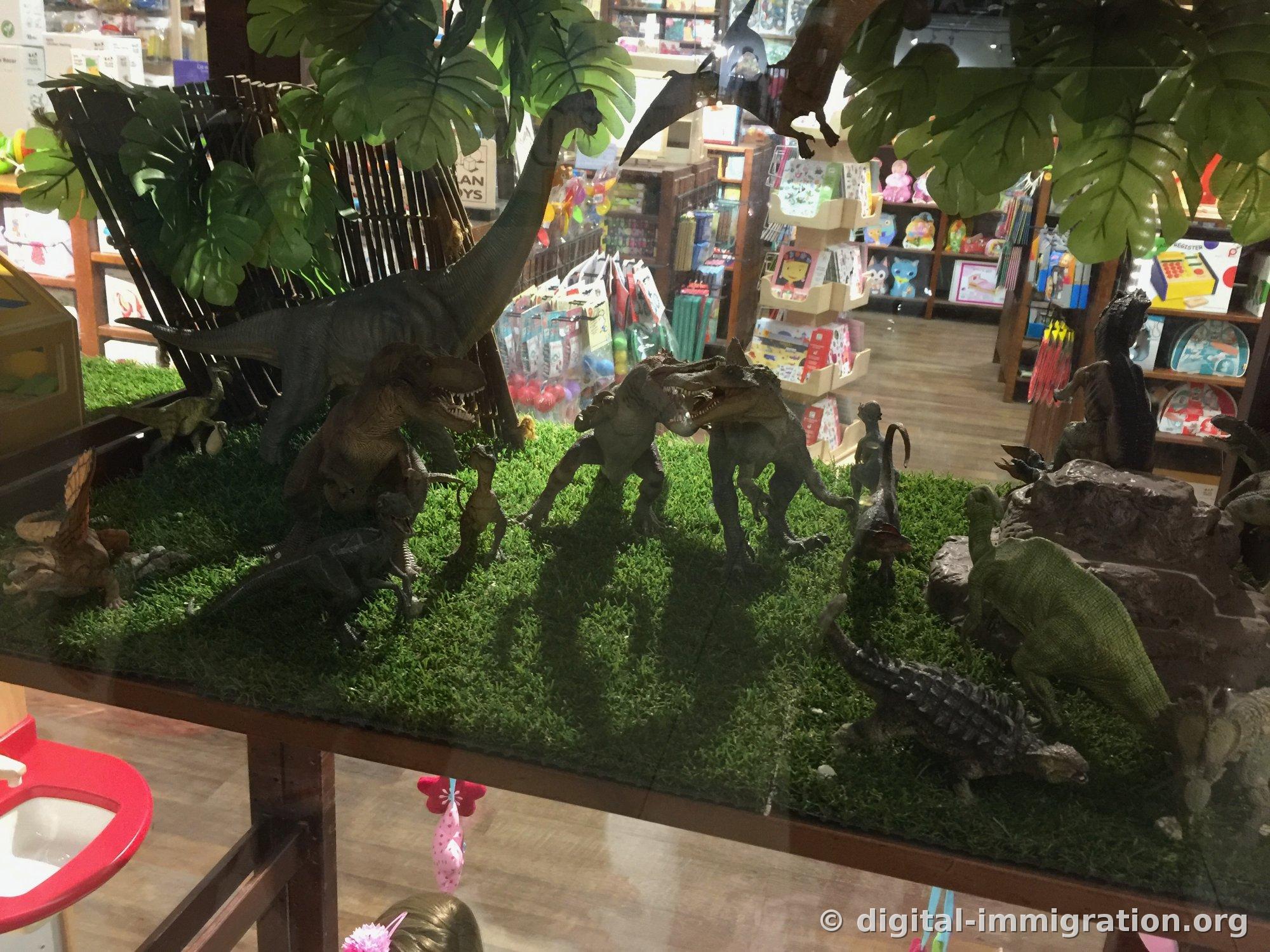 Un diorama de magasin de jouets avec un Brachiosaure, un Tyranosaure et un Ankylosaure disposés sur du gazon artificiel