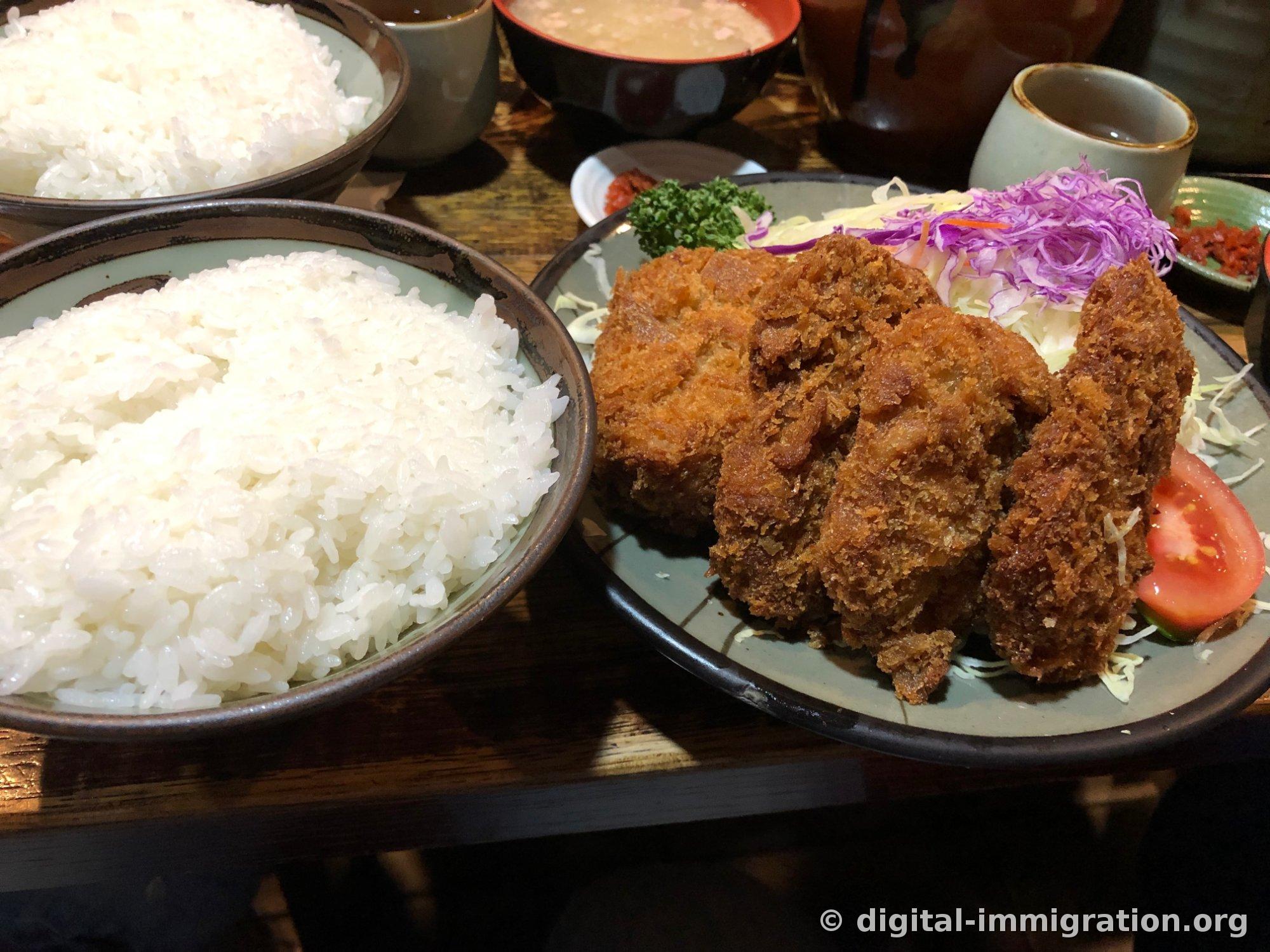 Two heaping bowls of white rice on a wooden table, alongside a gray plate with thick-sliced fried cutlet accompanied by shredded cabbage, red cabbage, and tomato, and a red lacquered miso soup