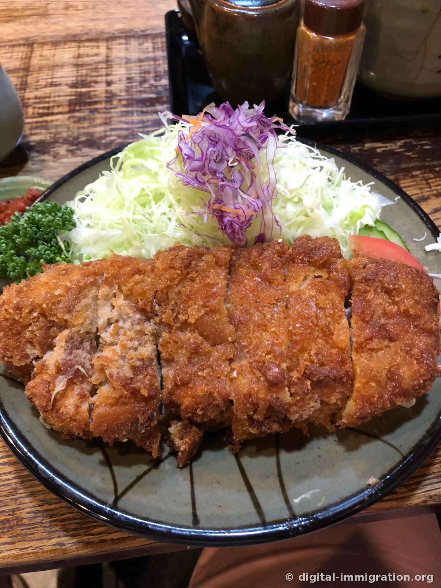 A thick-sliced, dark brown tonkatsu on a gray plate piled with shredded cabbage, with a bottle of red shichimi spice in the background on the condiment stand