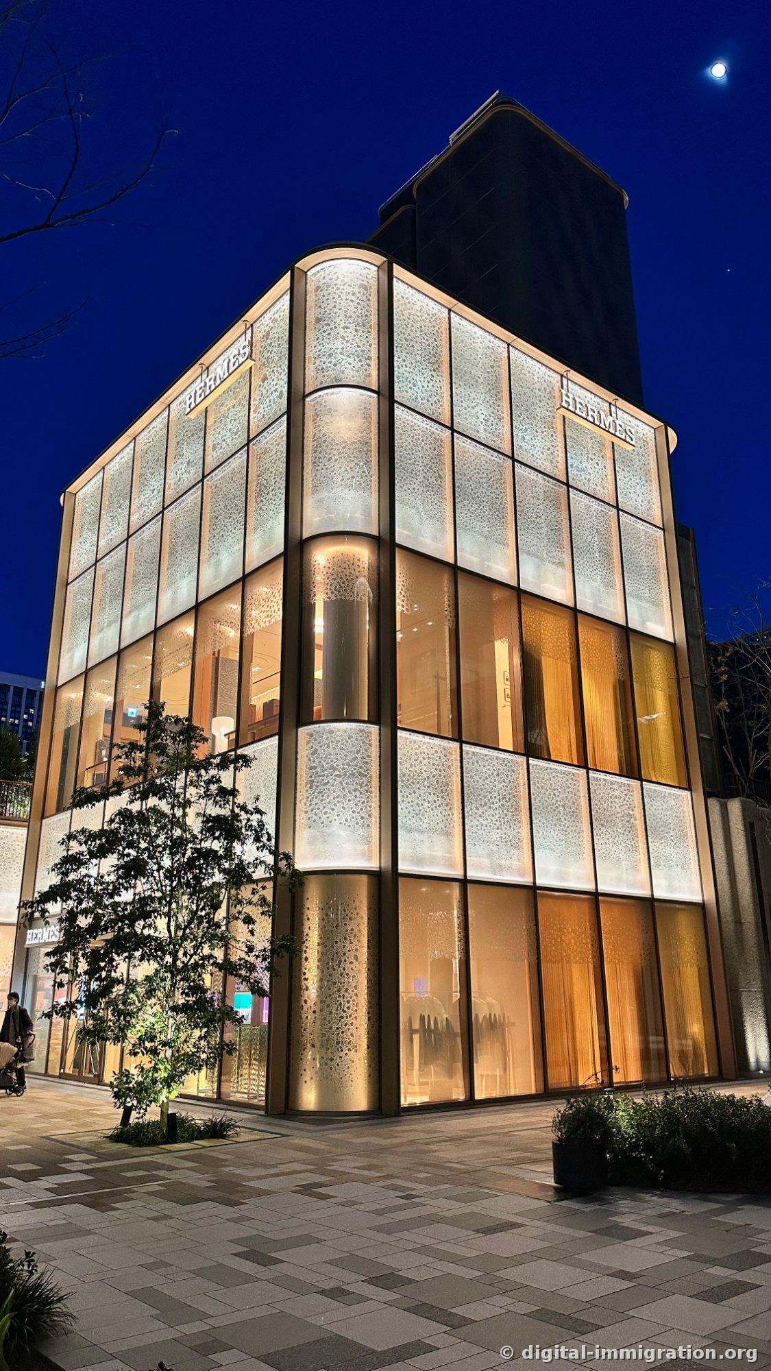 Corner view of the illuminated Hermès Azabudai store at night, with a crescent moon above the glass facade