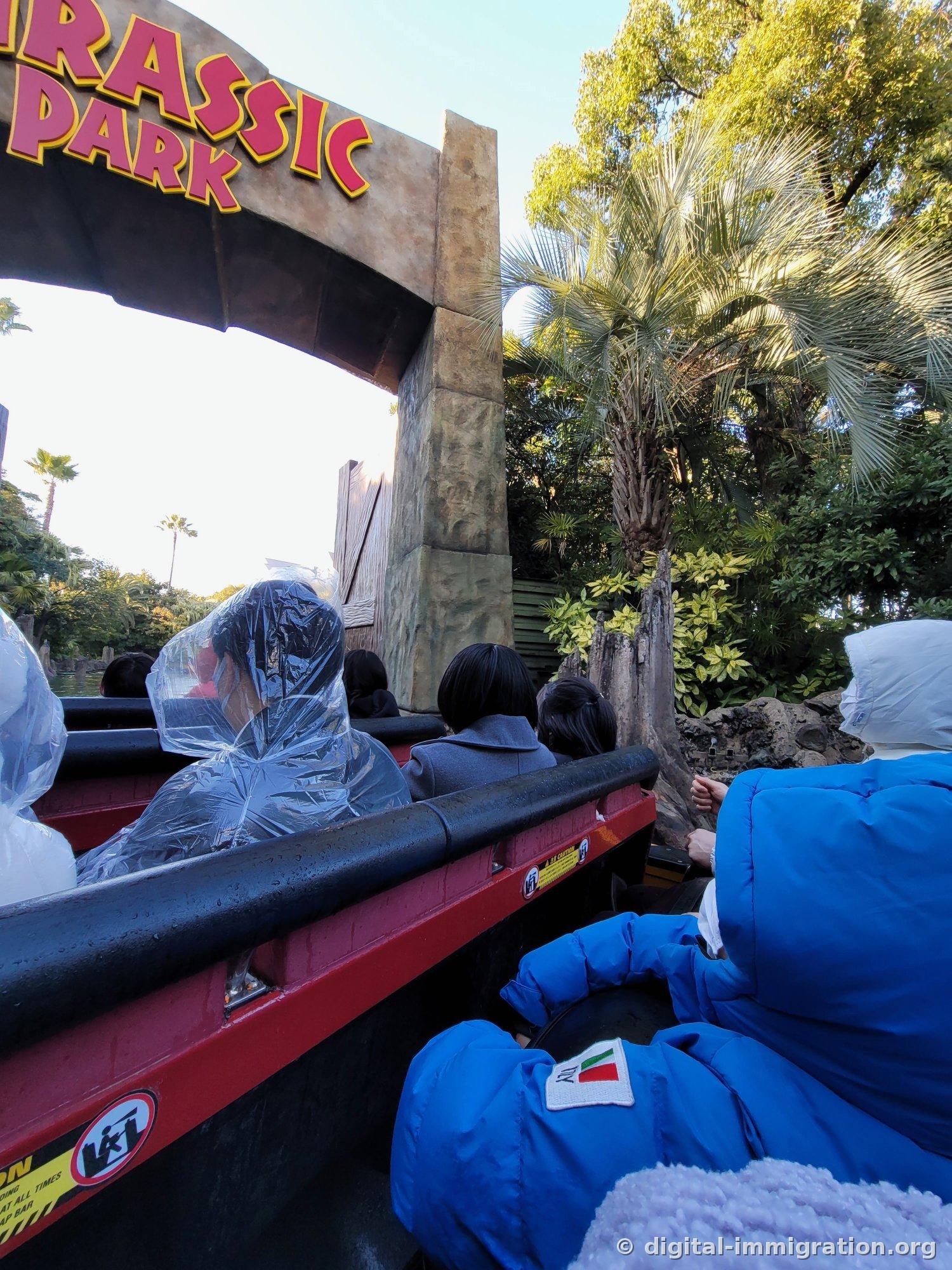 Red boat passing under the Jurassic Park sign with passengers wearing plastic ponchos, jungle palm scenery visible