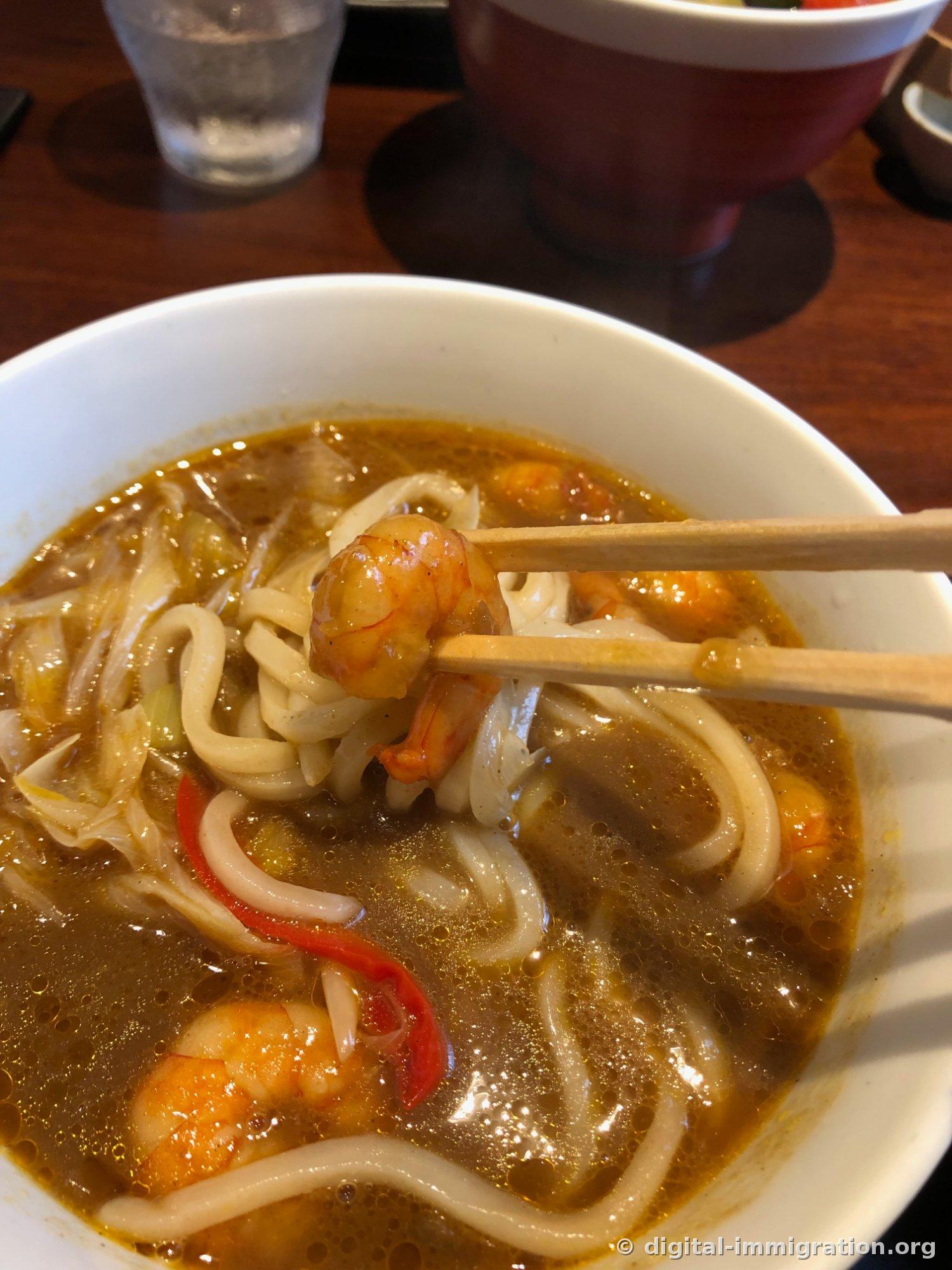 箸でカレースープの中の殻付きえび一尾を持ち上げたクローズアップ。スープの下に赤い唐辛子の破片が見える