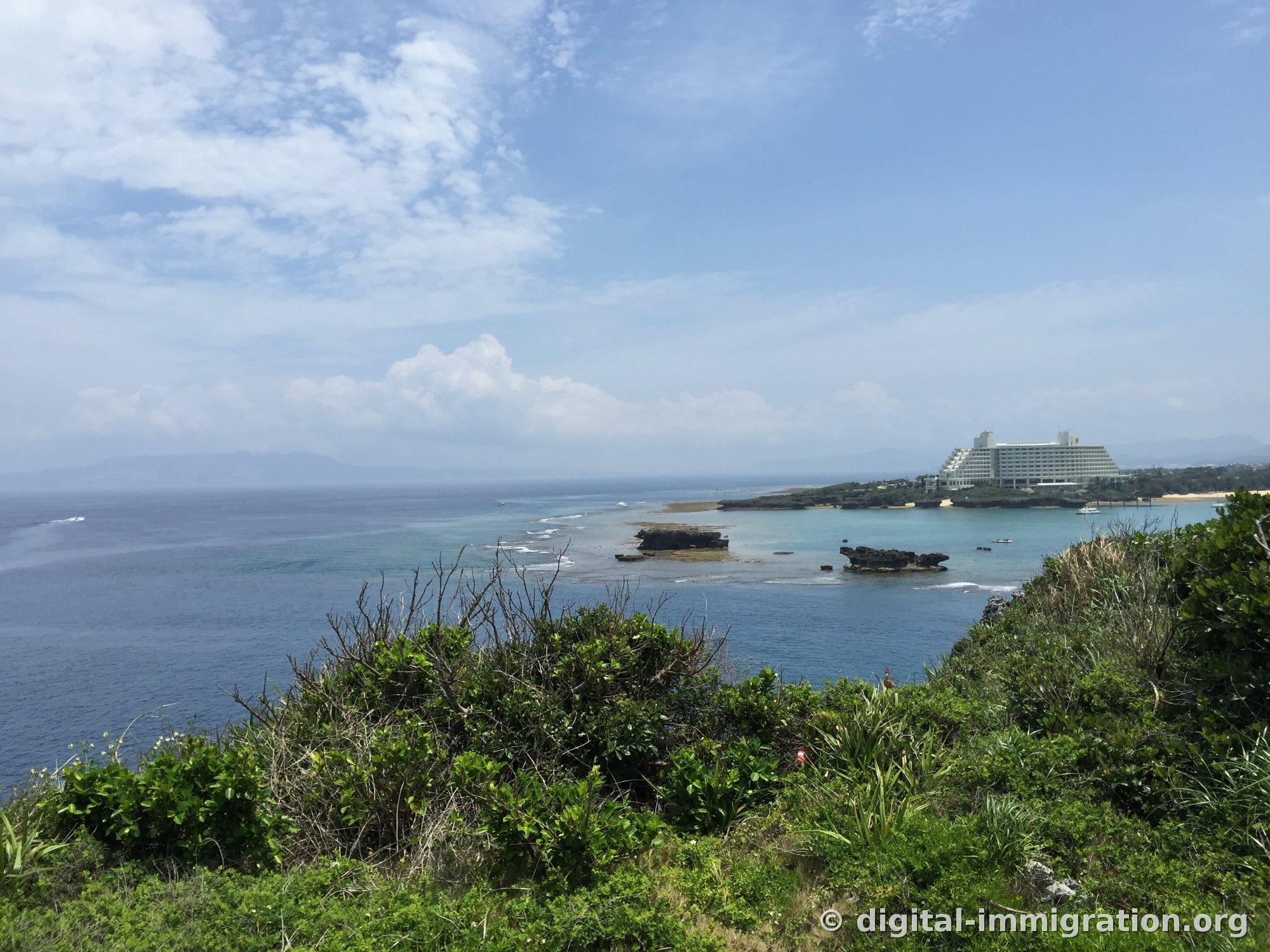 灌木の茂み越しに広がる沖縄西海岸。サンゴ礁の上で波が砕け、遠く岬の上に階段式のリゾートホテルが立っている