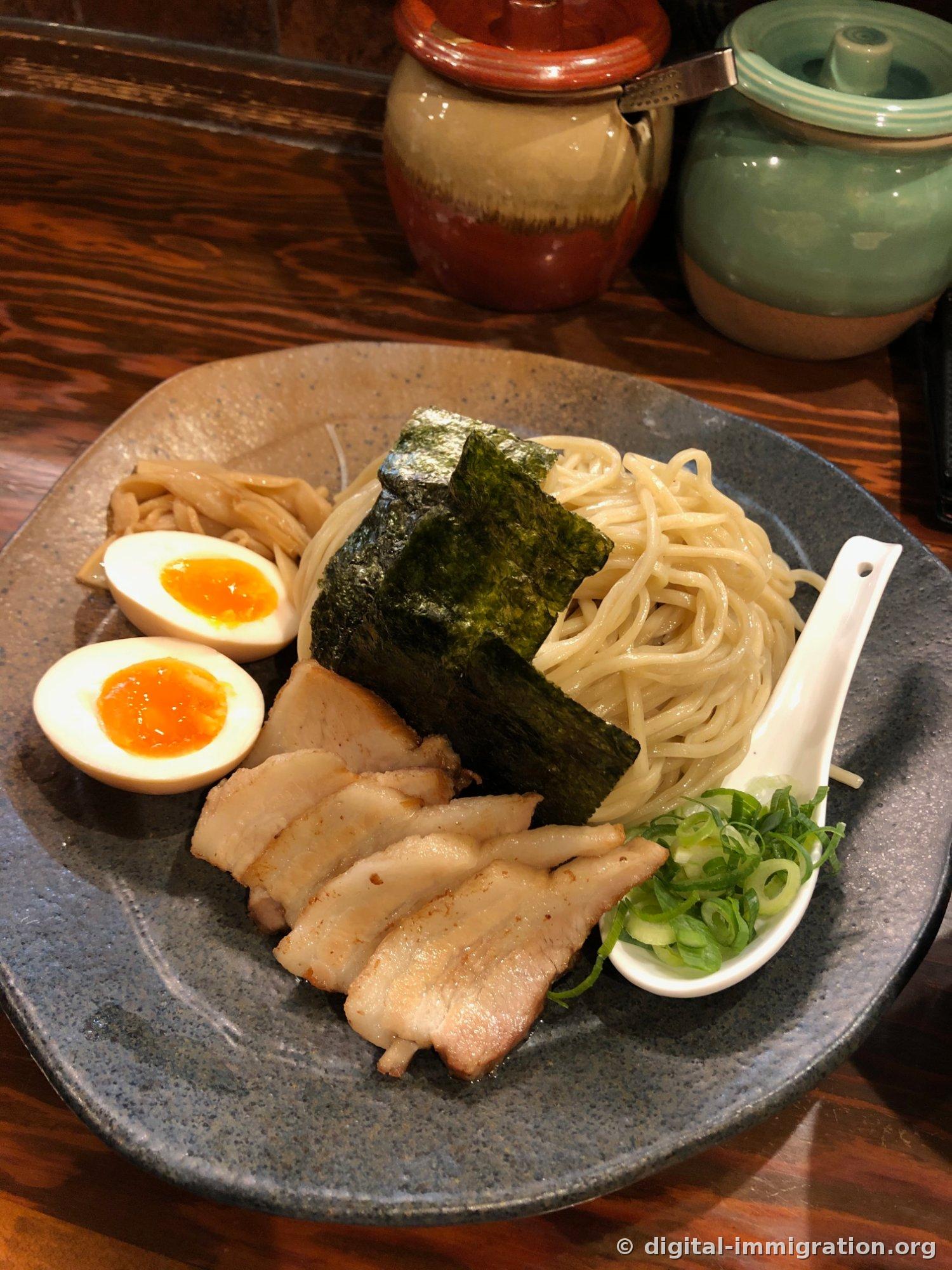 Le tsukemen servi sur une assiette en céramique grise. Une grande feuille d'algue nori au-dessus des nouilles, du menma et deux moitiés d'œuf mi-cuit à gauche, quatre tranches de char siu au centre, oignons verts hachés dans un petit bol blanc à droite